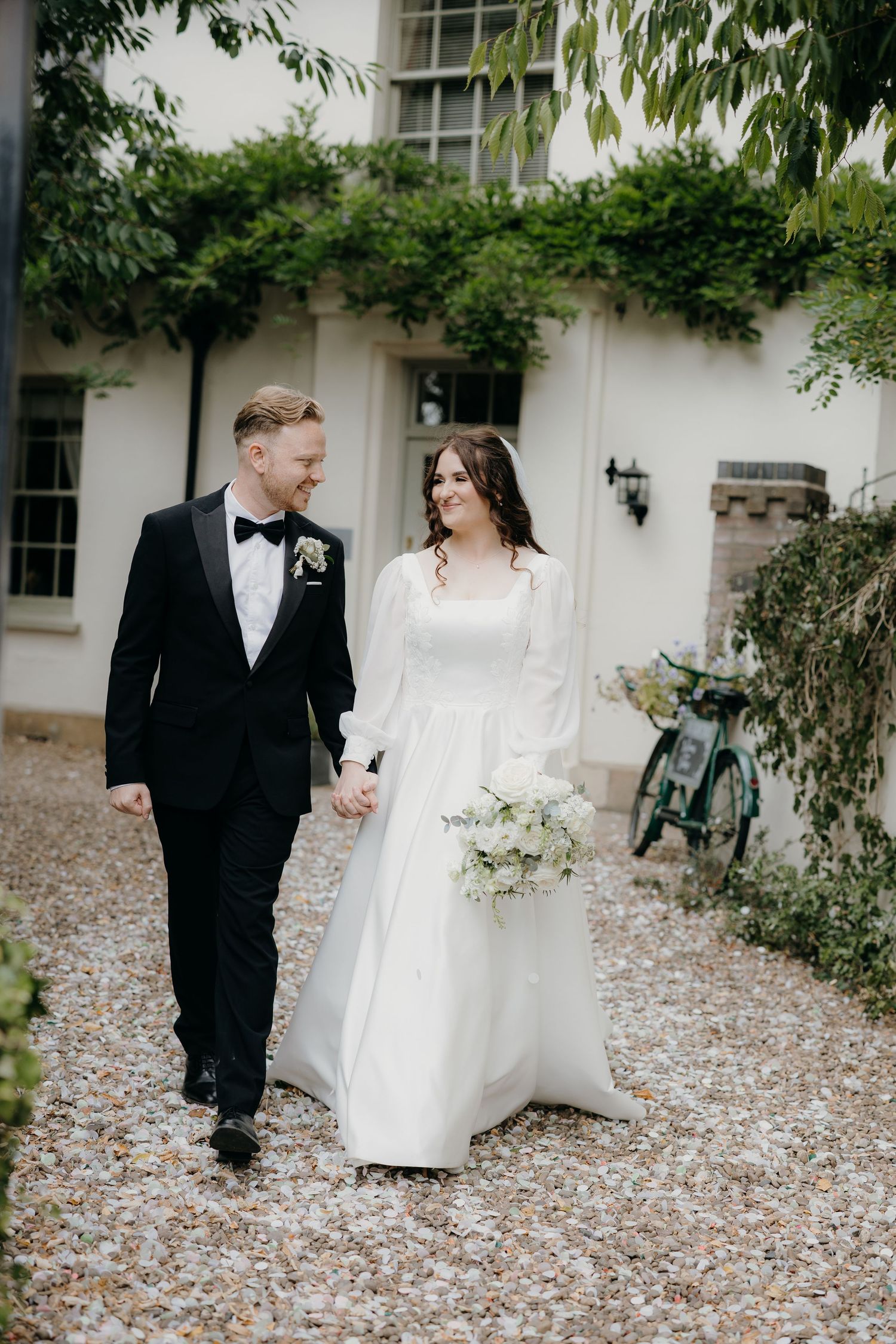 Bride and groom walk hand-in-hand on gravel path, she holds white bouquet, he wears black tuxedo, ivy-covered building behind.