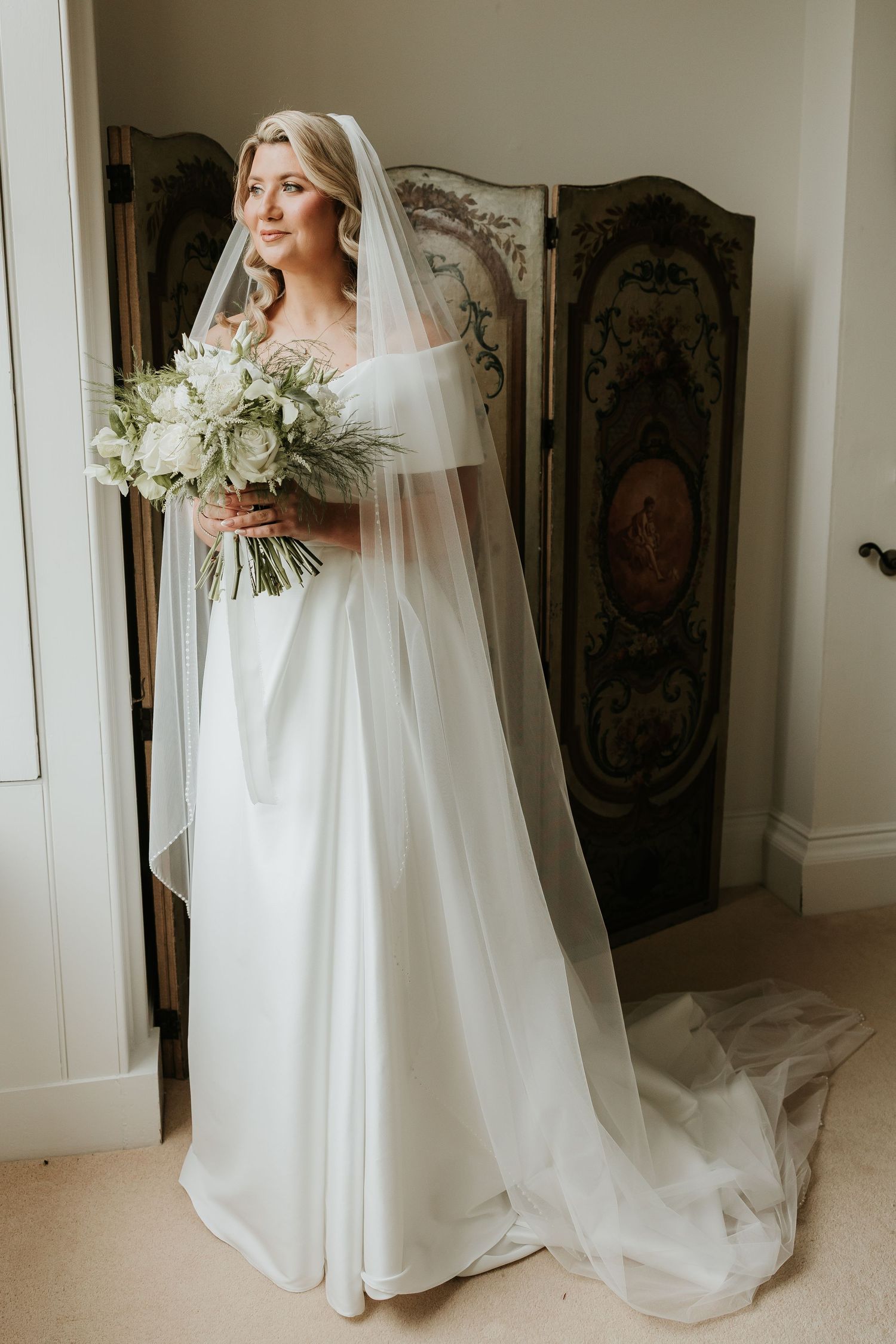 Bride in an off-the-shoulder white satin wedding gown and long sheer veil holding a white floral bouquet beside an ornate folding screen.
