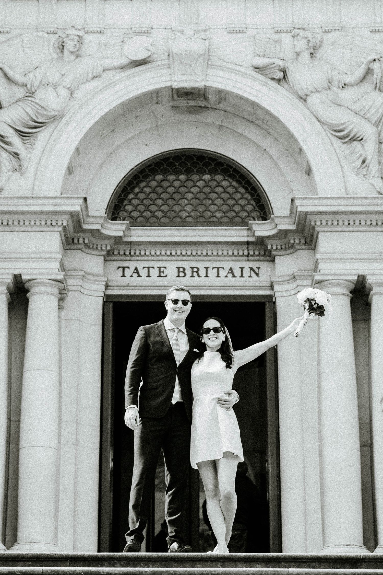 Black and white photo of a couple posing on the steps of Tate Britain, London, with the woman holding a bouquet.