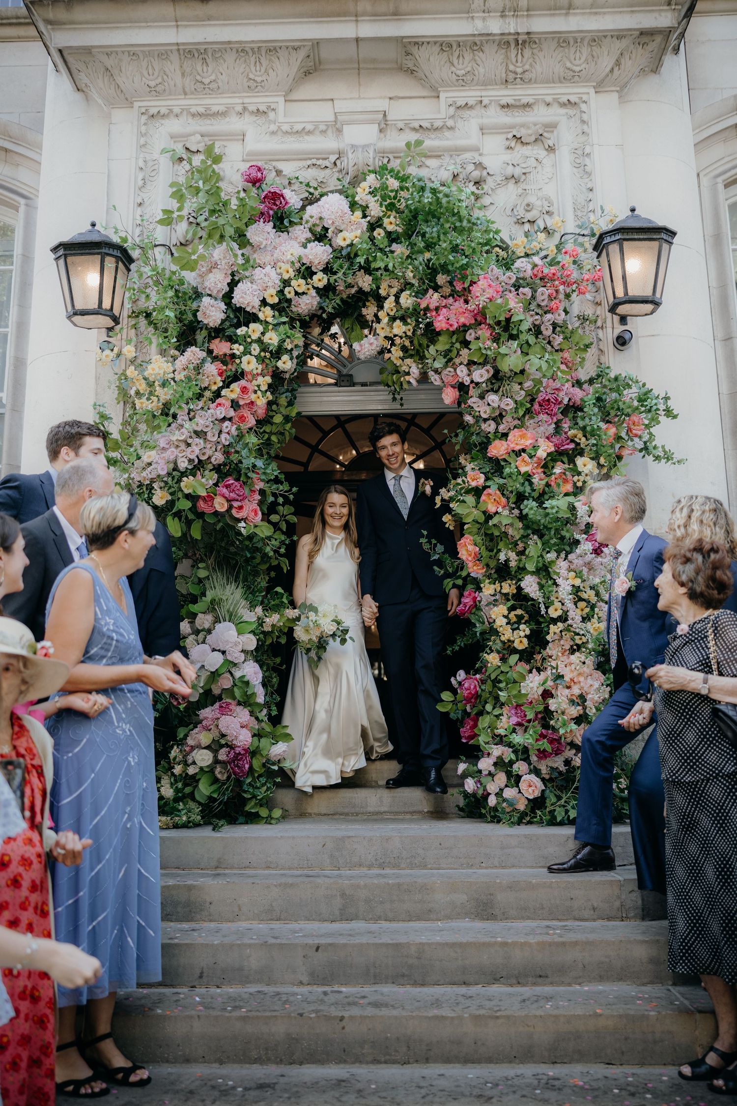 Bride and groom stand in a doorway beneath a lush pink floral arch as wedding guests line the stone steps and watch.