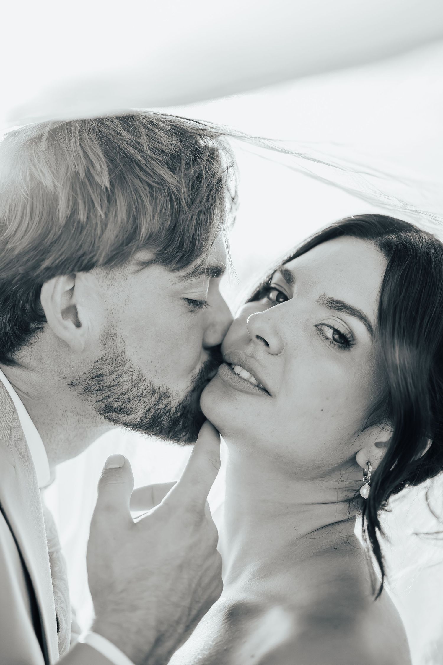 Black and white wedding portrait of a bearded groom kissing the cheek of a smiling bride in a sheer veil with an updo and drop earring.