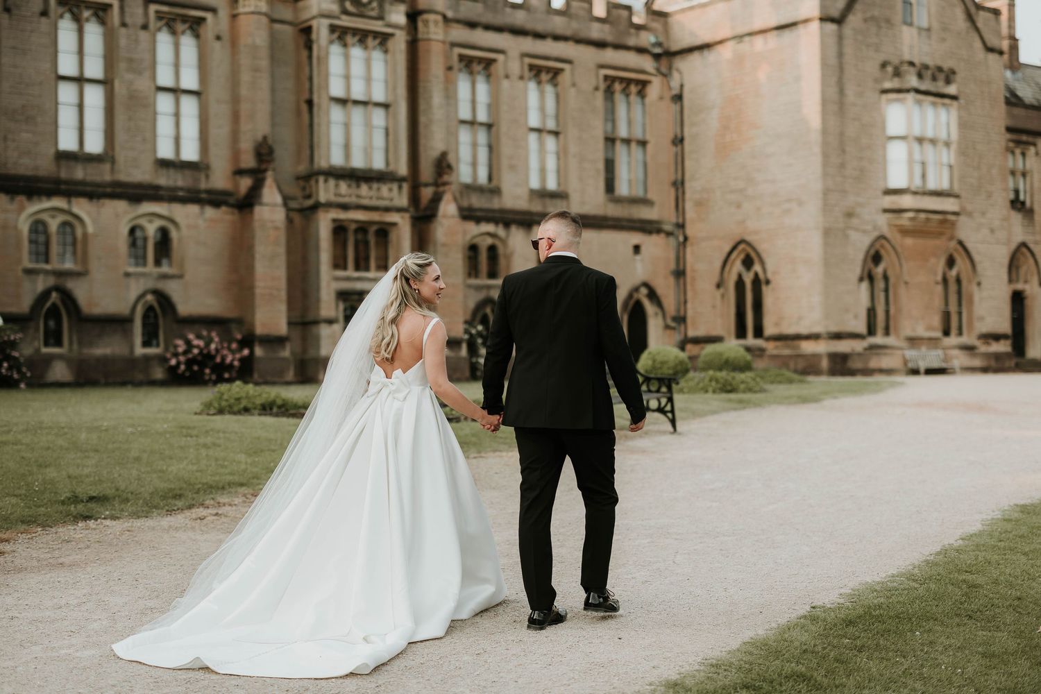 Bride and groom walk hand-in-hand towards Newstead Abbey, bride in elegant backless gown with flowing veil.