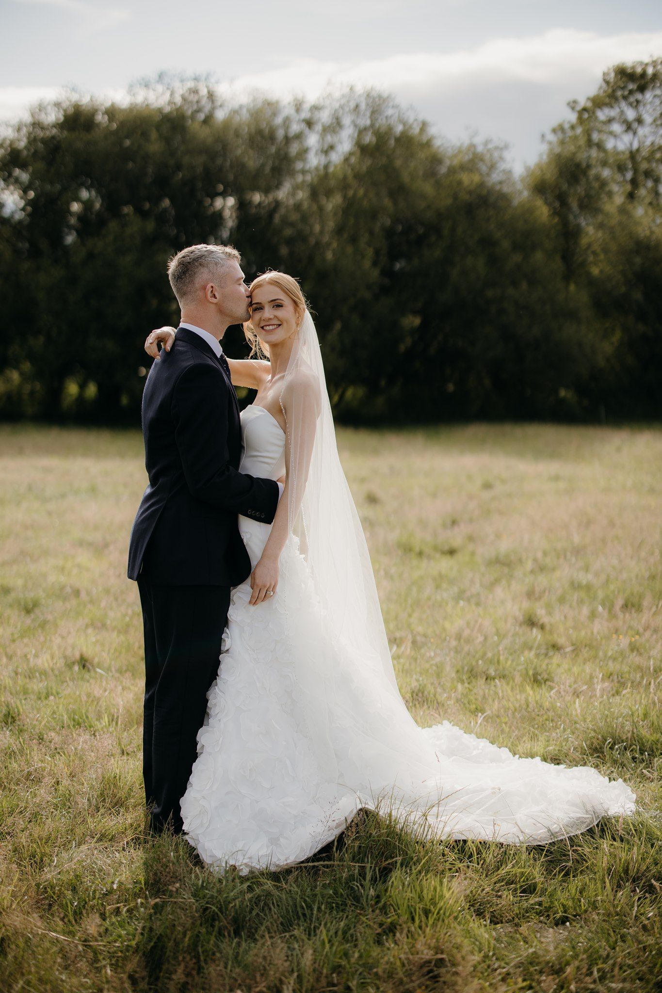 A bride and groom embrace in a lush green field, she wearing a white gown with veil, he in a dark suit.