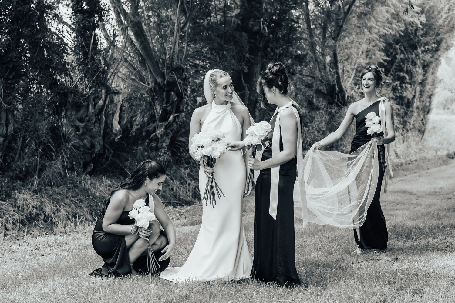 A black and white wedding photo showing preparations outdoors with bridal party members helping adjust the dress.