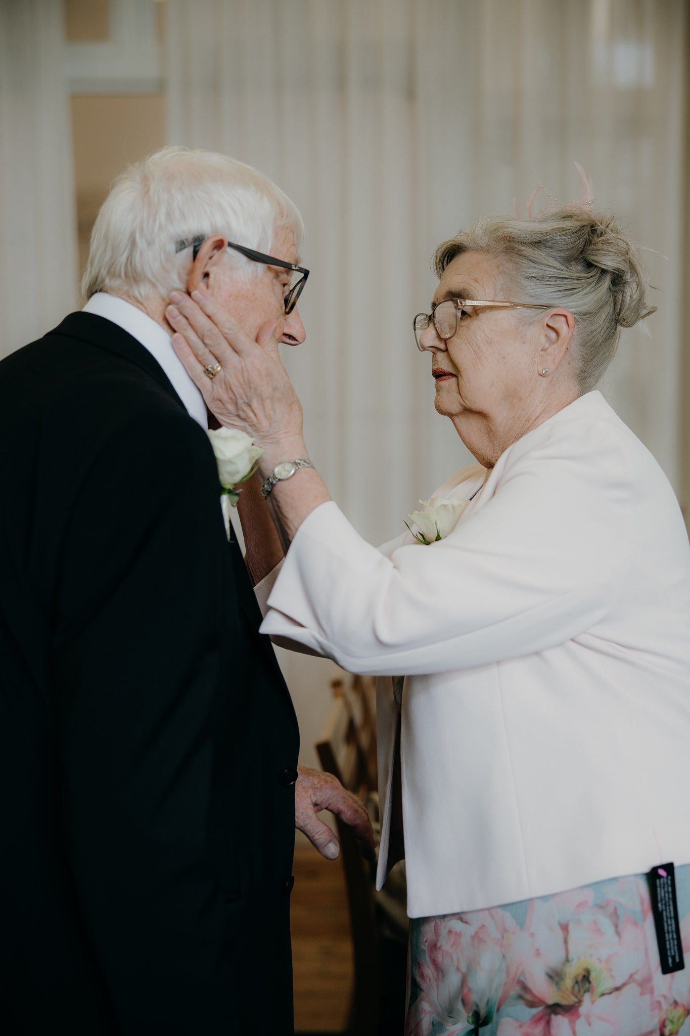 Elderly couple sharing a tender moment, woman gently touching man's face, both wearing white boutonnieres.