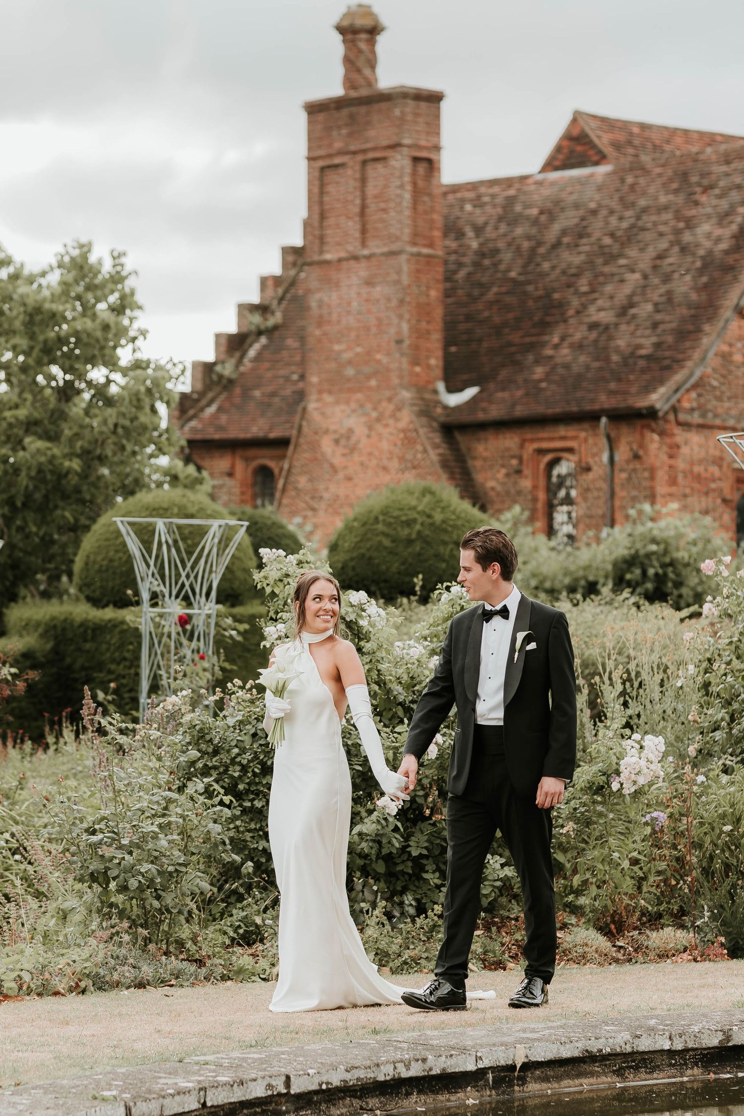 Couple enjoys romantic moment walking along garden path by historic brick mansion with ornate chimney on overcast day.