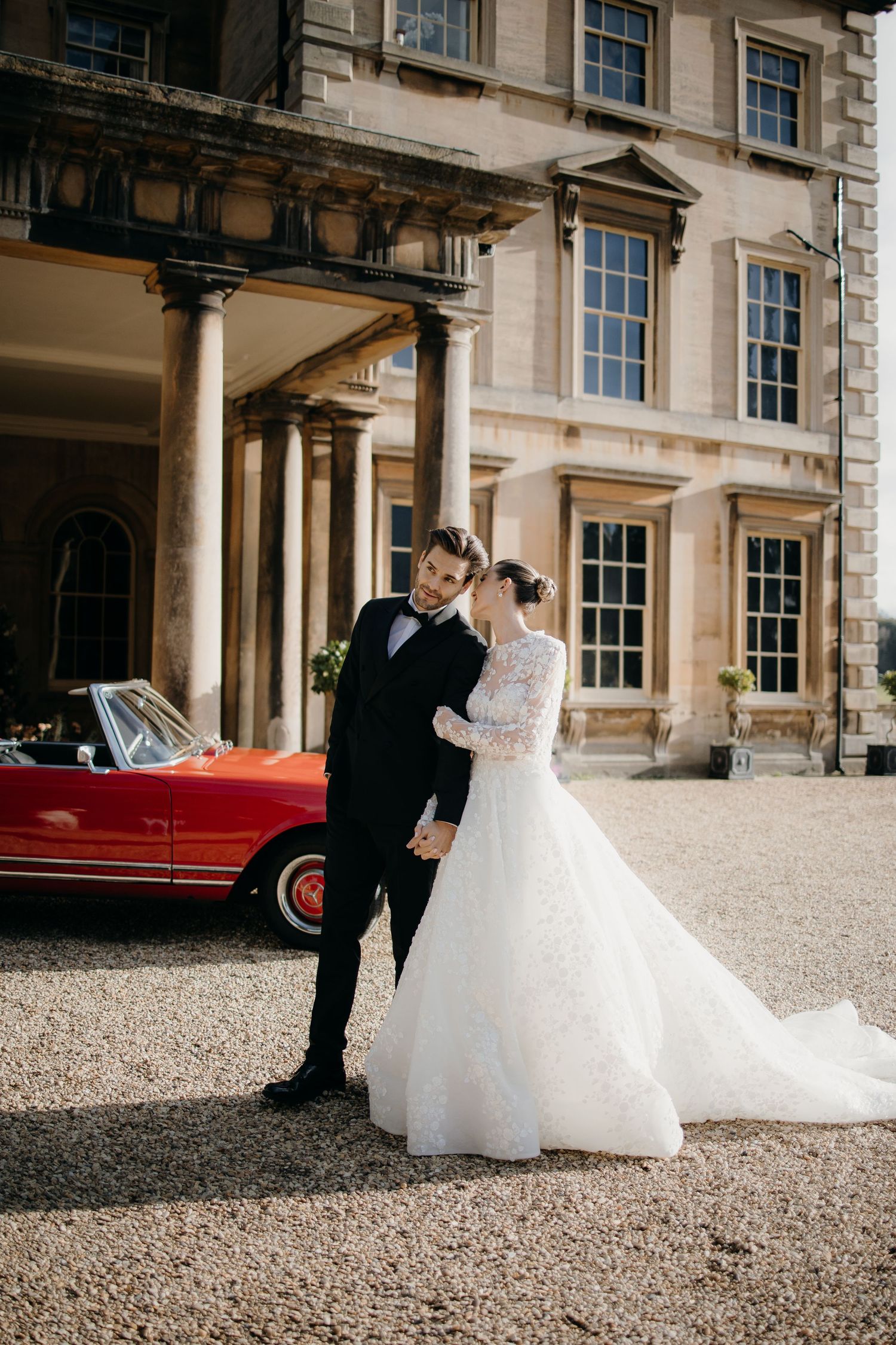 Bride and groom embrace in front of Prestwold hall with a red vintage convertible car on a gravel driveway.