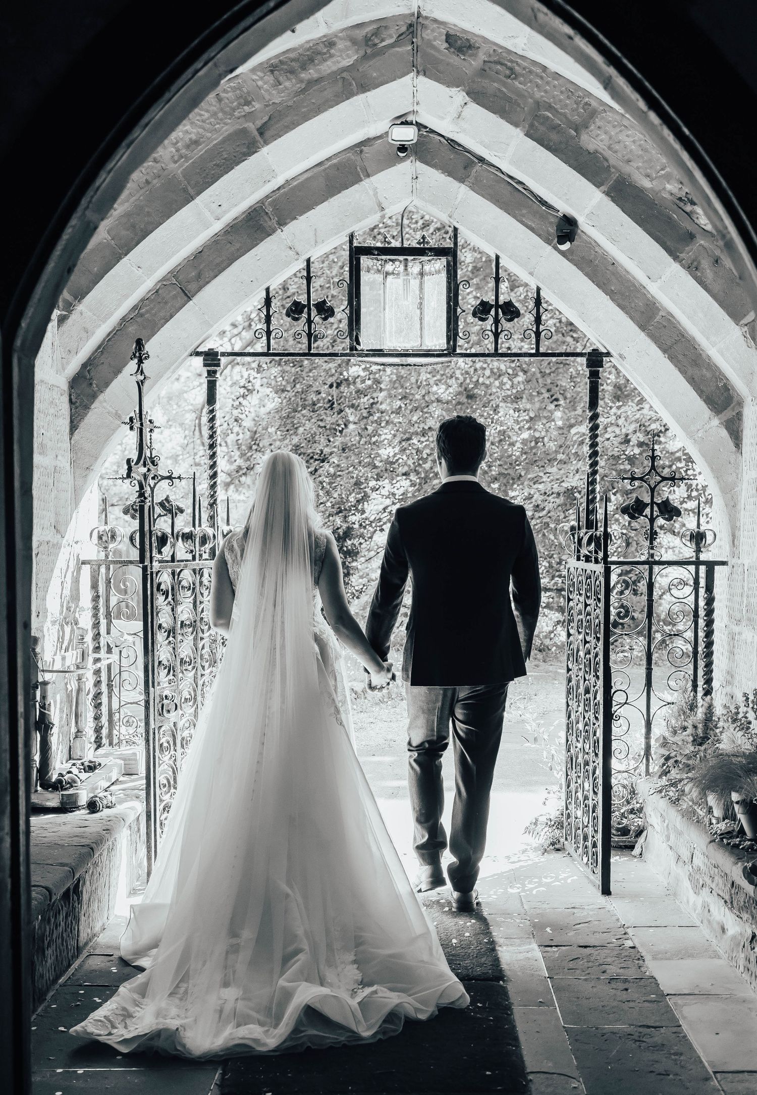 A bride and groom stand in silhouette under a stone church archway decorated with ornate lanterns and wrought iron gates.