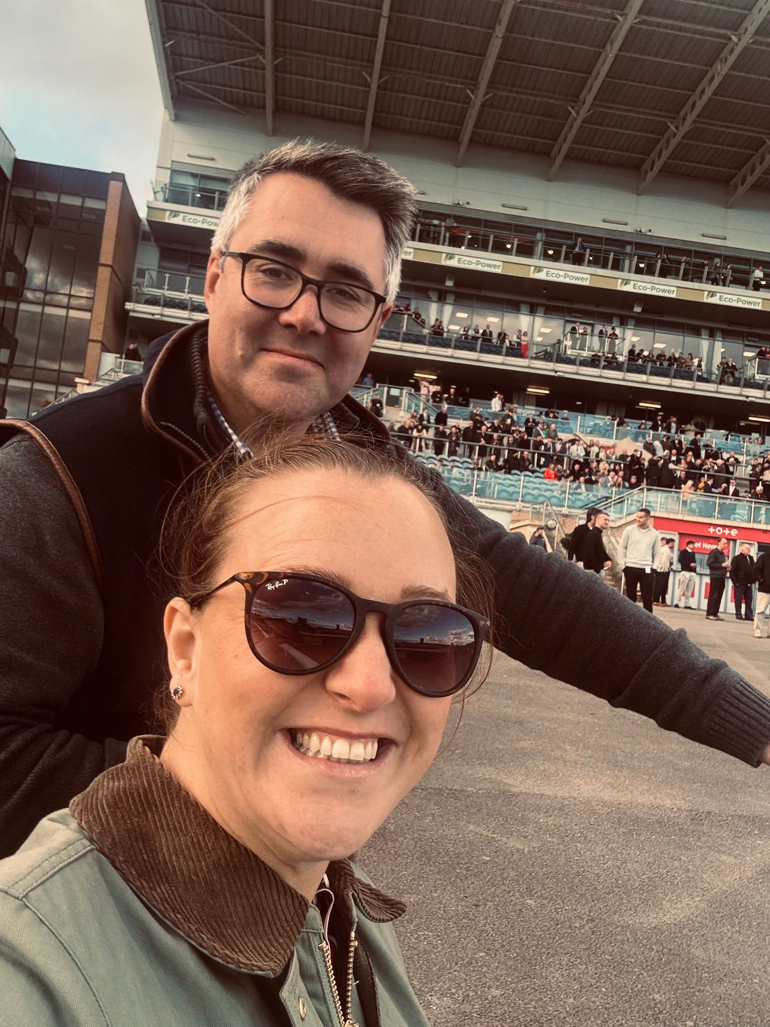 A selfie of two people wearing sunglasses and turtlenecks at a sports stadium with crowded stands visible in background.