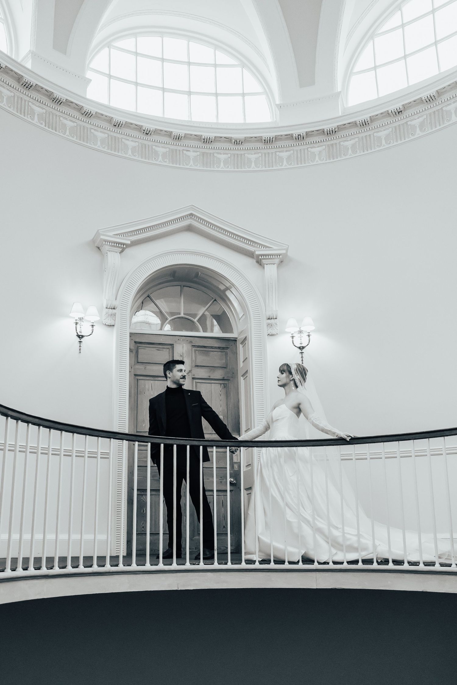 Bride in a flowing white gown and groom in a black suit hold hands on a curved balcony beneath an arched doorway in an elegant venue.