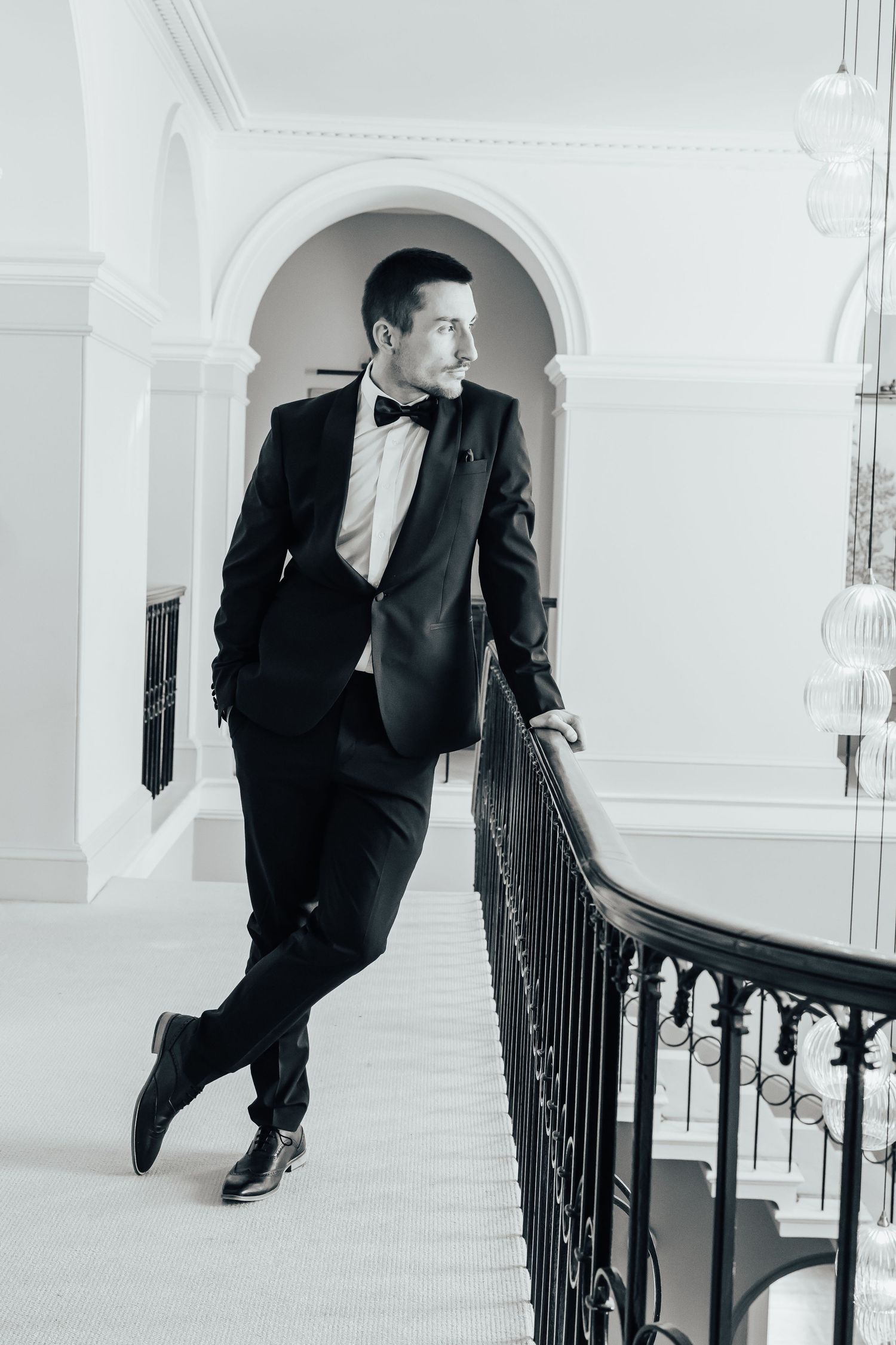 Elegant man in tuxedo and bow tie leaning on staircase railing in a grand white interior, black and white photo.