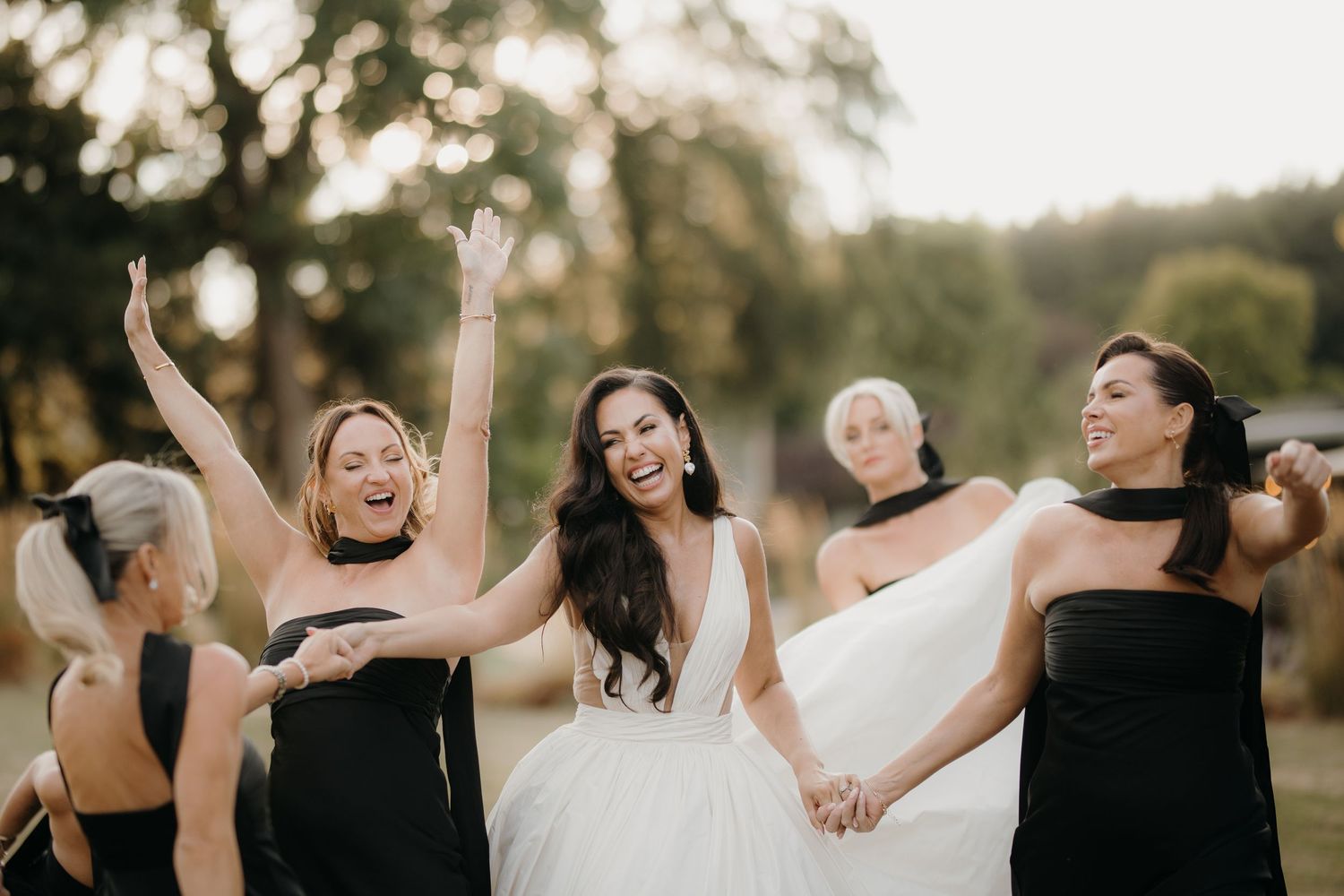 Laughing bride in a white wedding gown holds hands with bridesmaids in black dresses as they celebrate outdoors among trees.