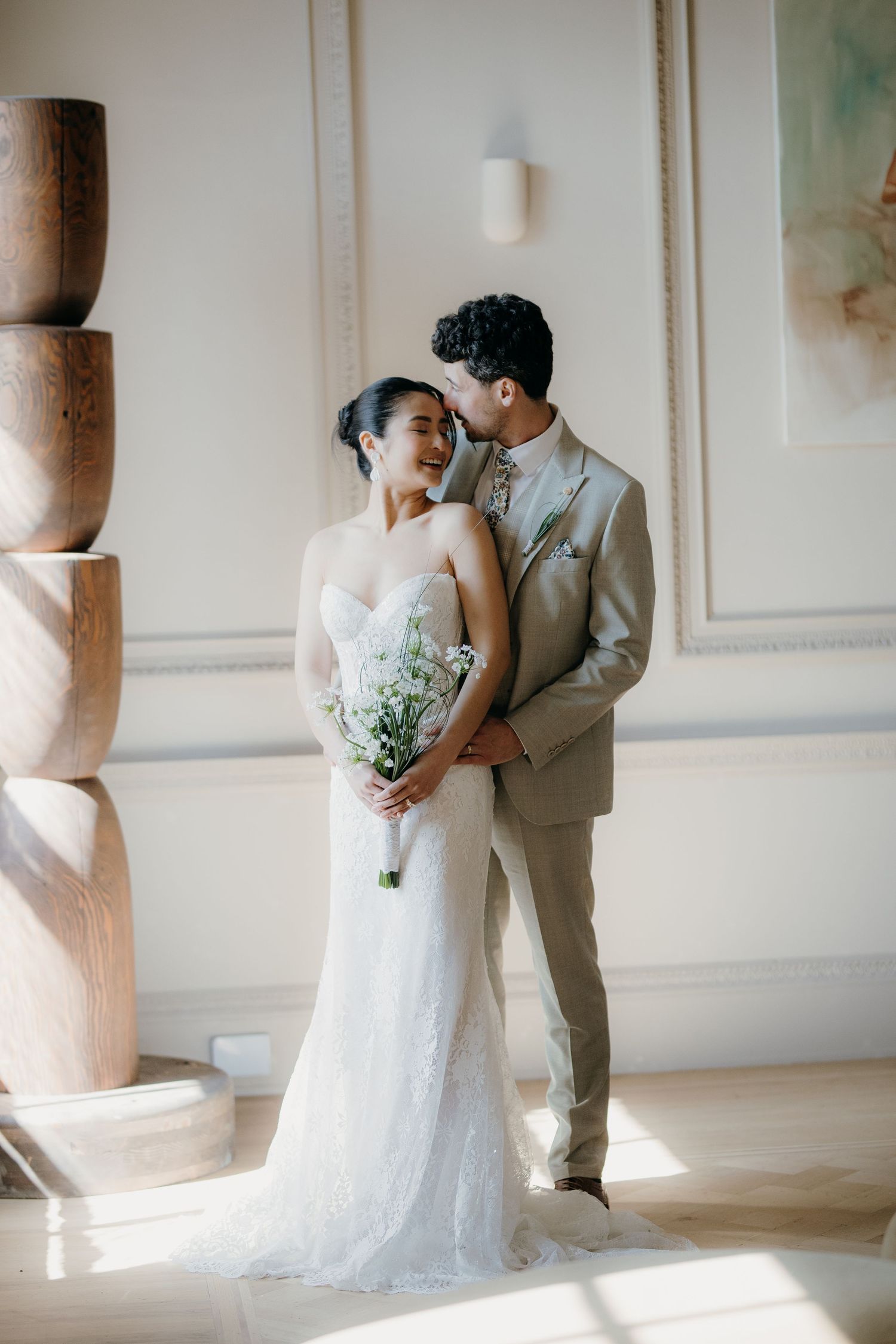 Bride in white lace gown holding floral bouquet embraced by groom in tan suit in elegant sunlit room at Denton Hall.