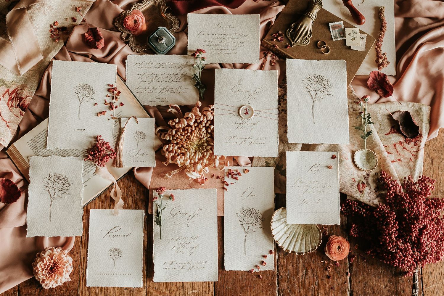 Wedding invitations and stationery laid out with dried flowers and blush pink decor on a wooden surface.
