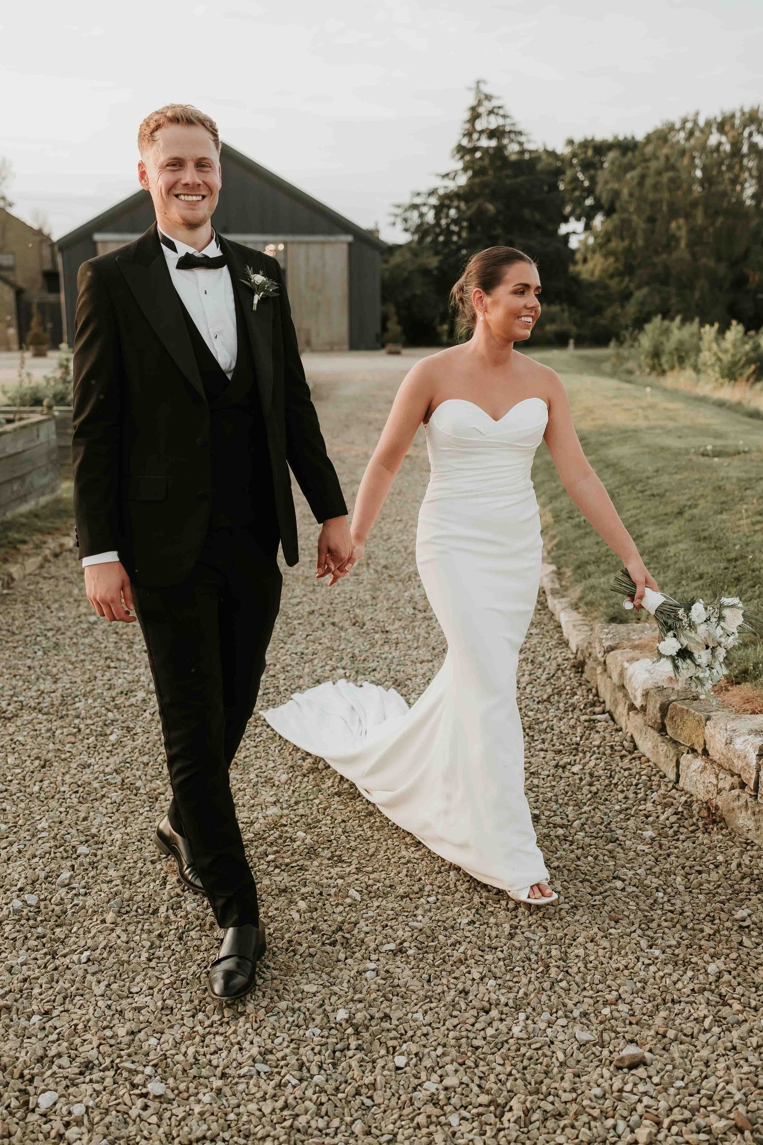 A couple in wedding attire walking hand in hand on a gravel path by a rustic barn.