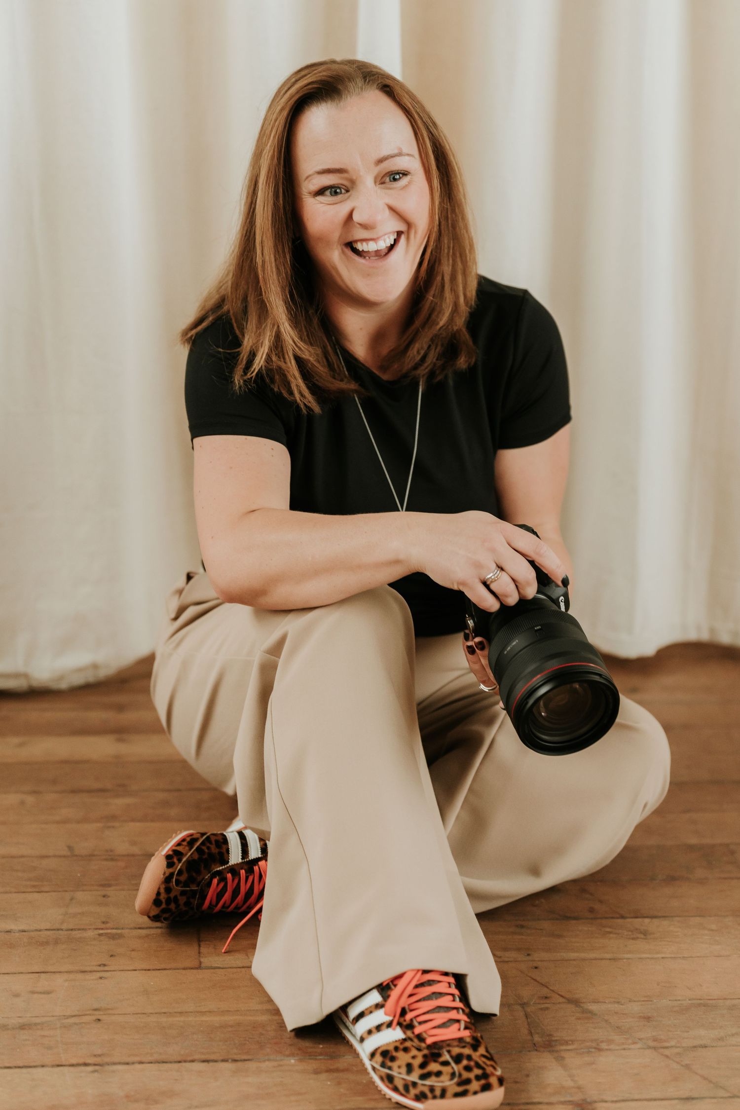 Smiling Sophie Ruck with red hair sitting on wood floor holding DSLR camera, wearing black top and khaki pants.
