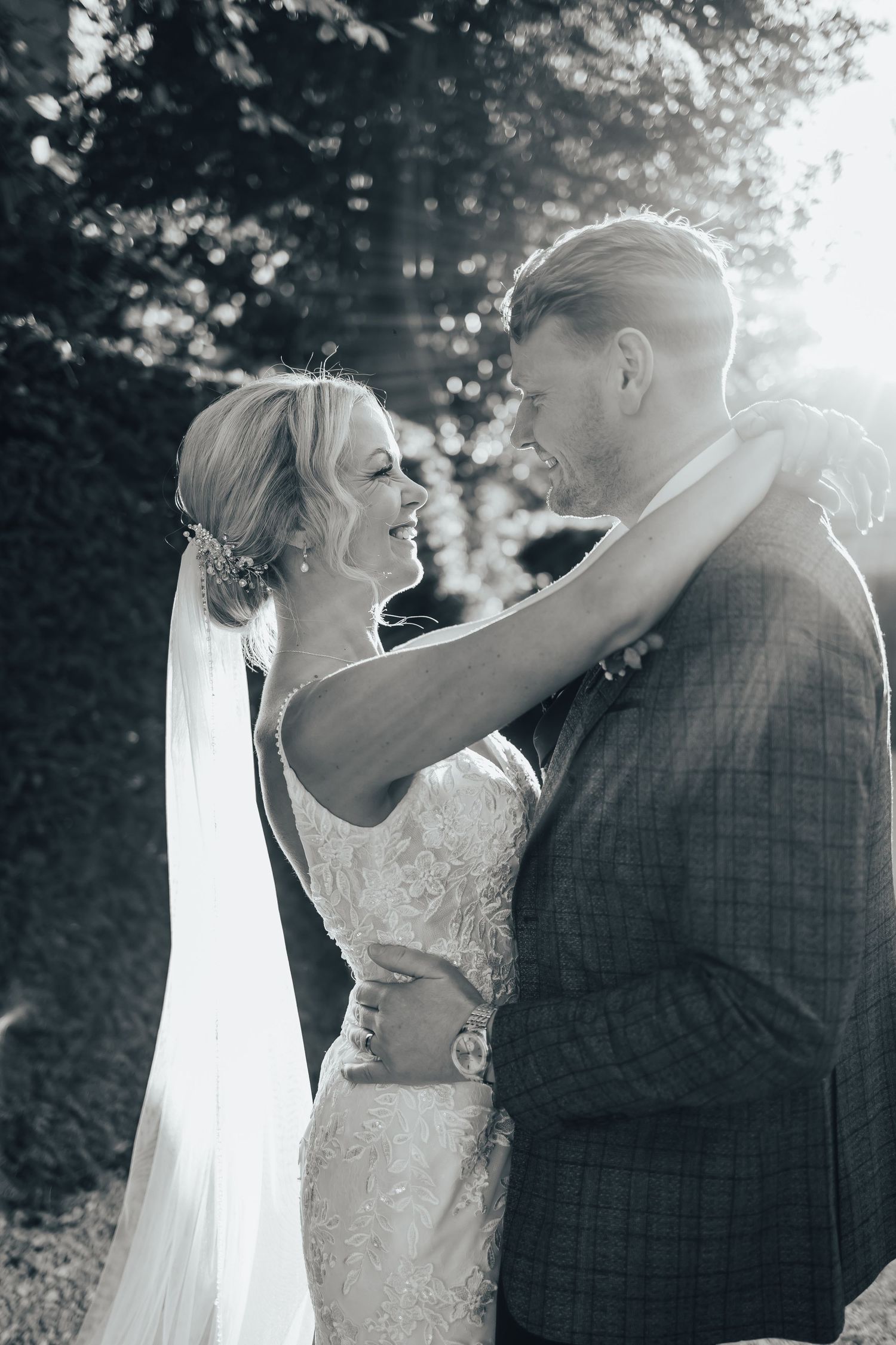 Black and white photo of a smiling bride and groom embracing outdoors with sunlight streaming through trees at Saltmarshe hall.
