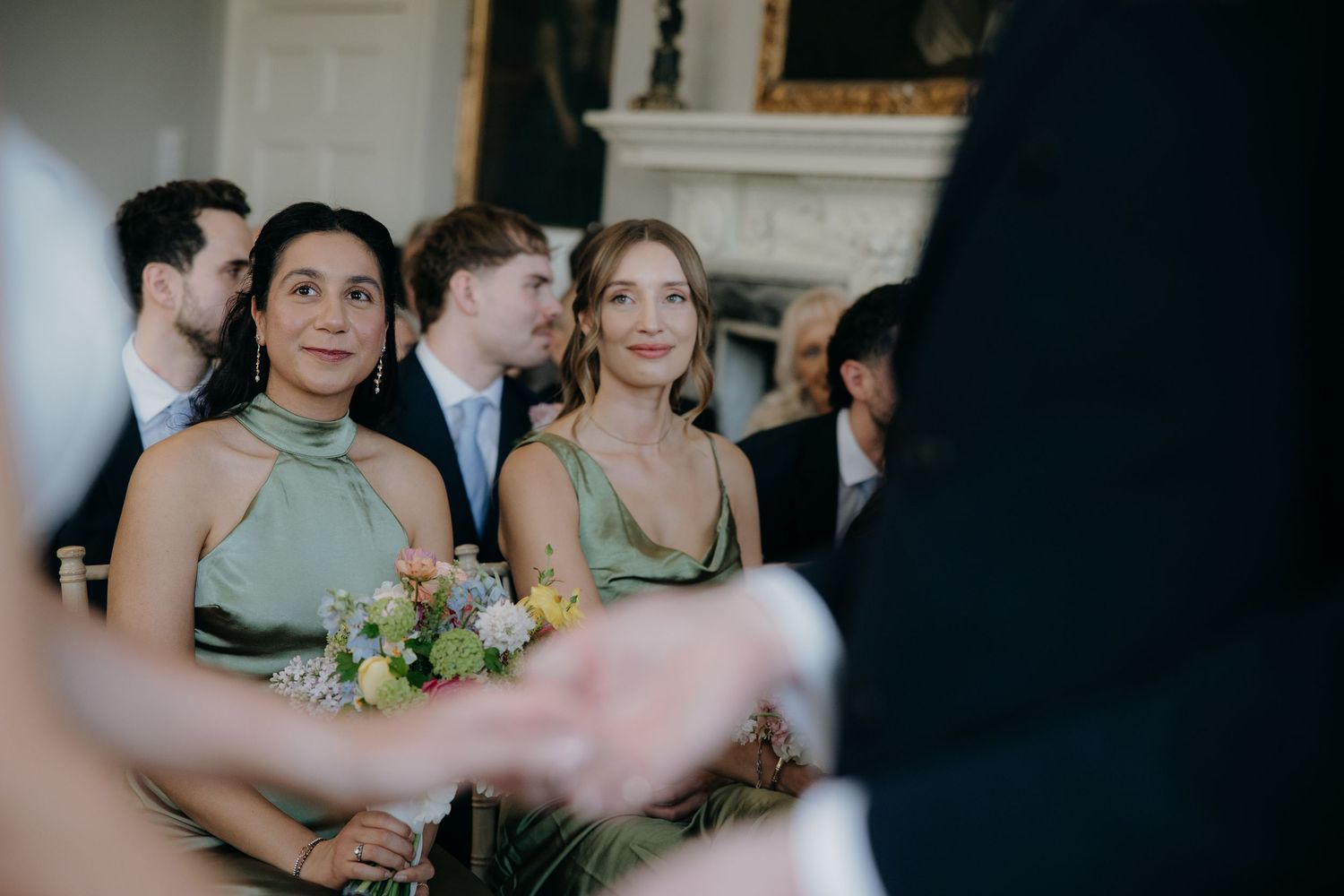 Two bridesmaids in sage green dresses smiling during a wedding ceremony in an elegant venue.