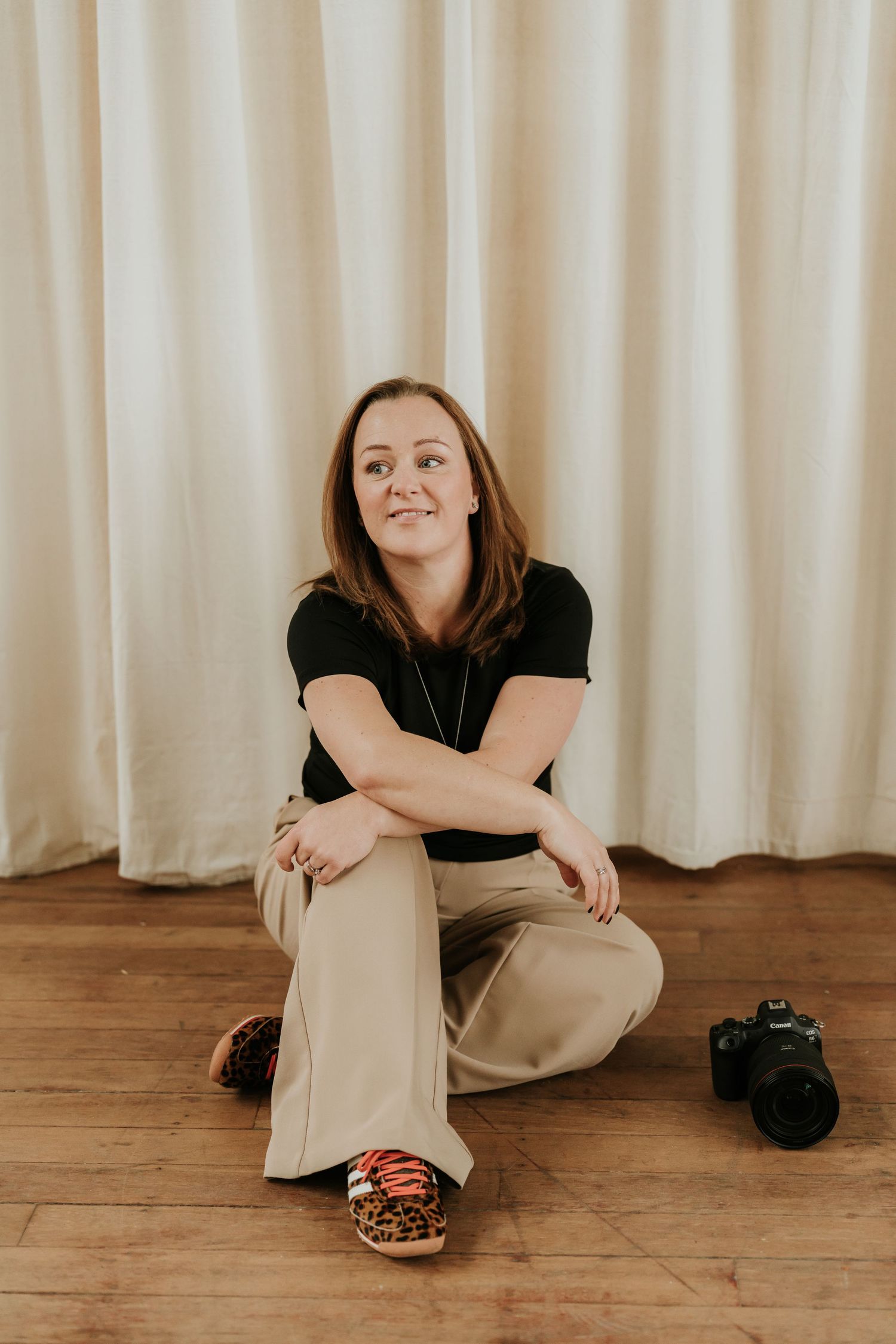 A photographer sits cross-legged on a wooden floor wearing a black shirt and khaki pants with a camera nearby.