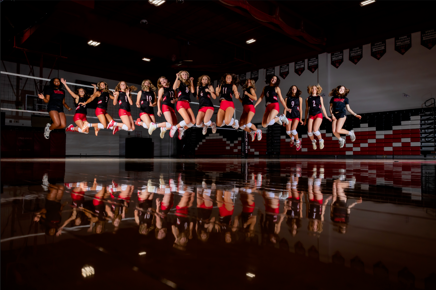 A cheerleading squad in red and black uniforms jumps in unison on a gym floor, with their reflection visible below.