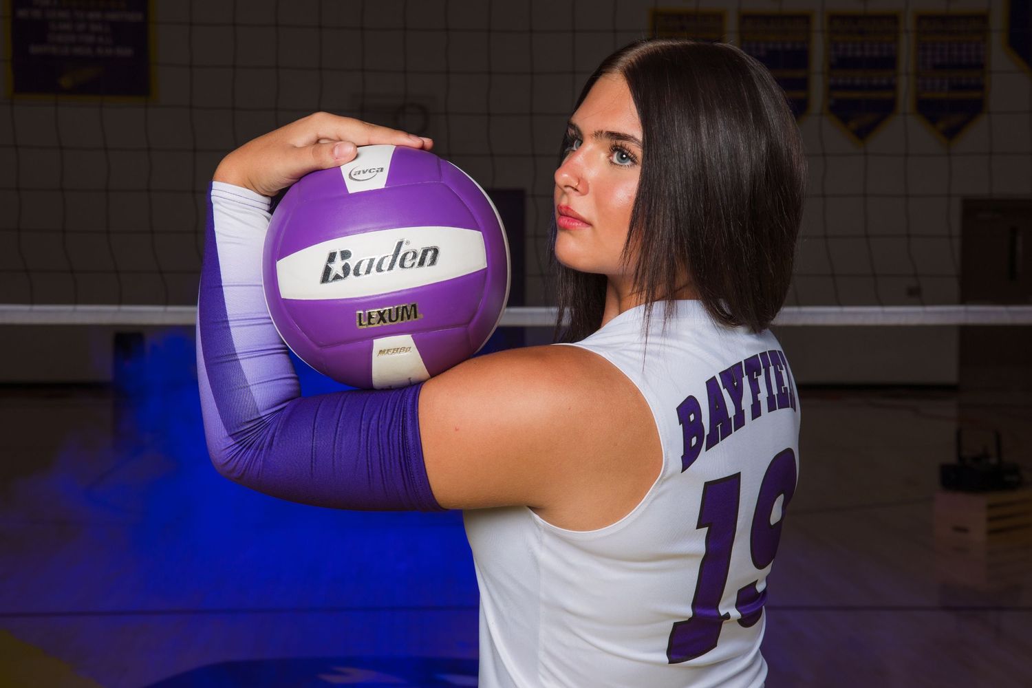 Volleyball player wearing white jersey prepares to serve during indoor match with purple ball.