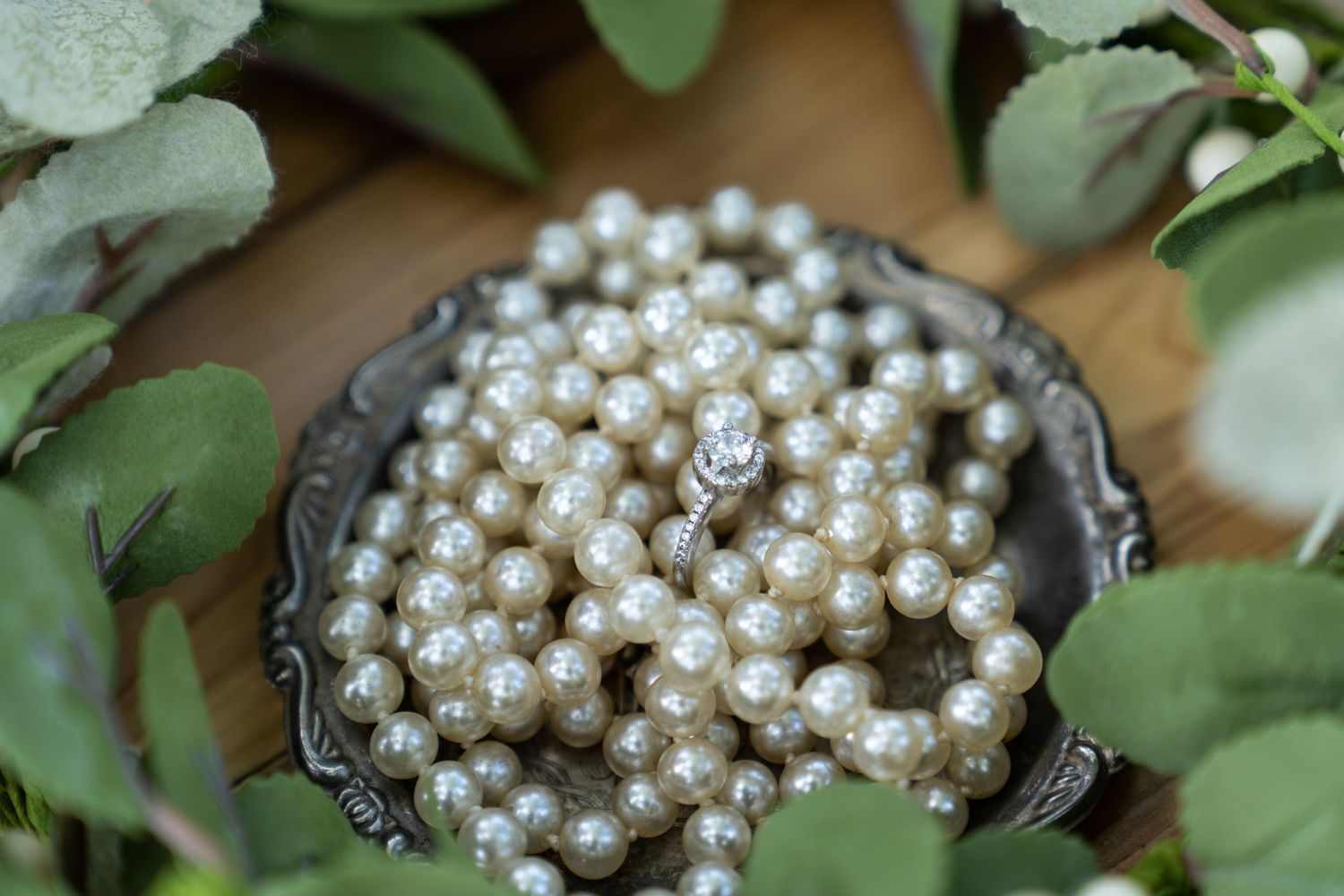 Brands photography of a diamond ring in a bowl of pearls surrounded by leaves.