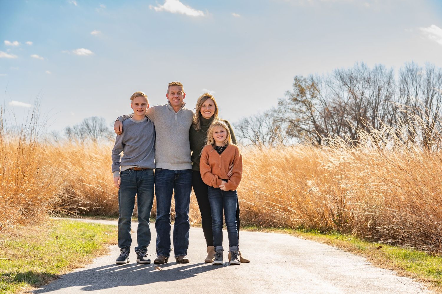 Fall family portrait of mom, dad, son and daughter standing on a wide walkway with tall grasses surrounding them.