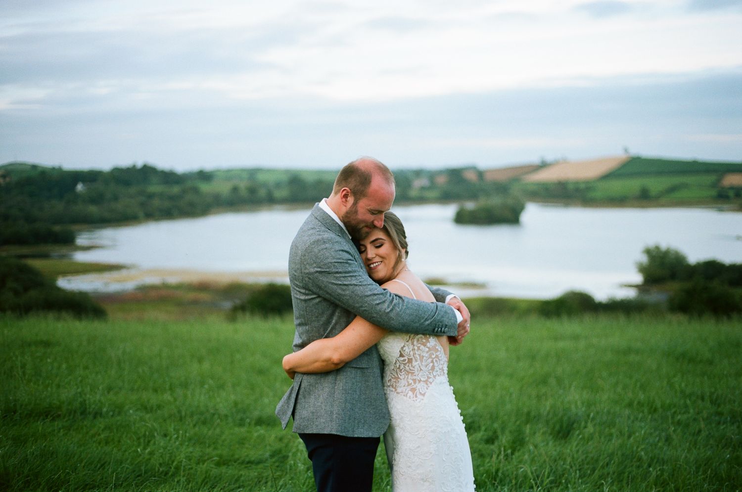 Newlyweds share an intimate moment on grassy hillside overlooking a serene lake.