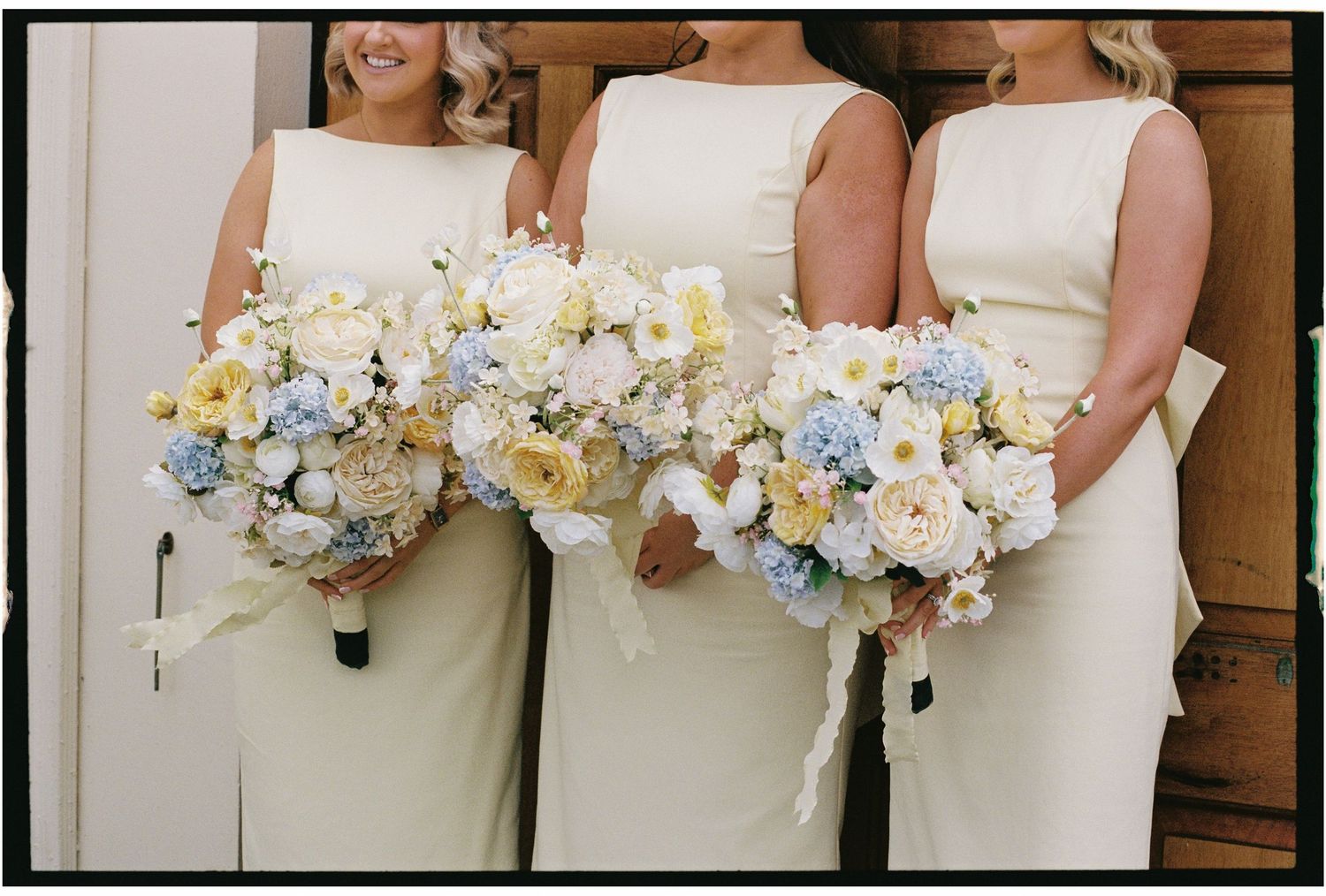 Three bridesmaids in white dresses hold elegant bouquets of white, yellow and blue flowers against a wooden wall.