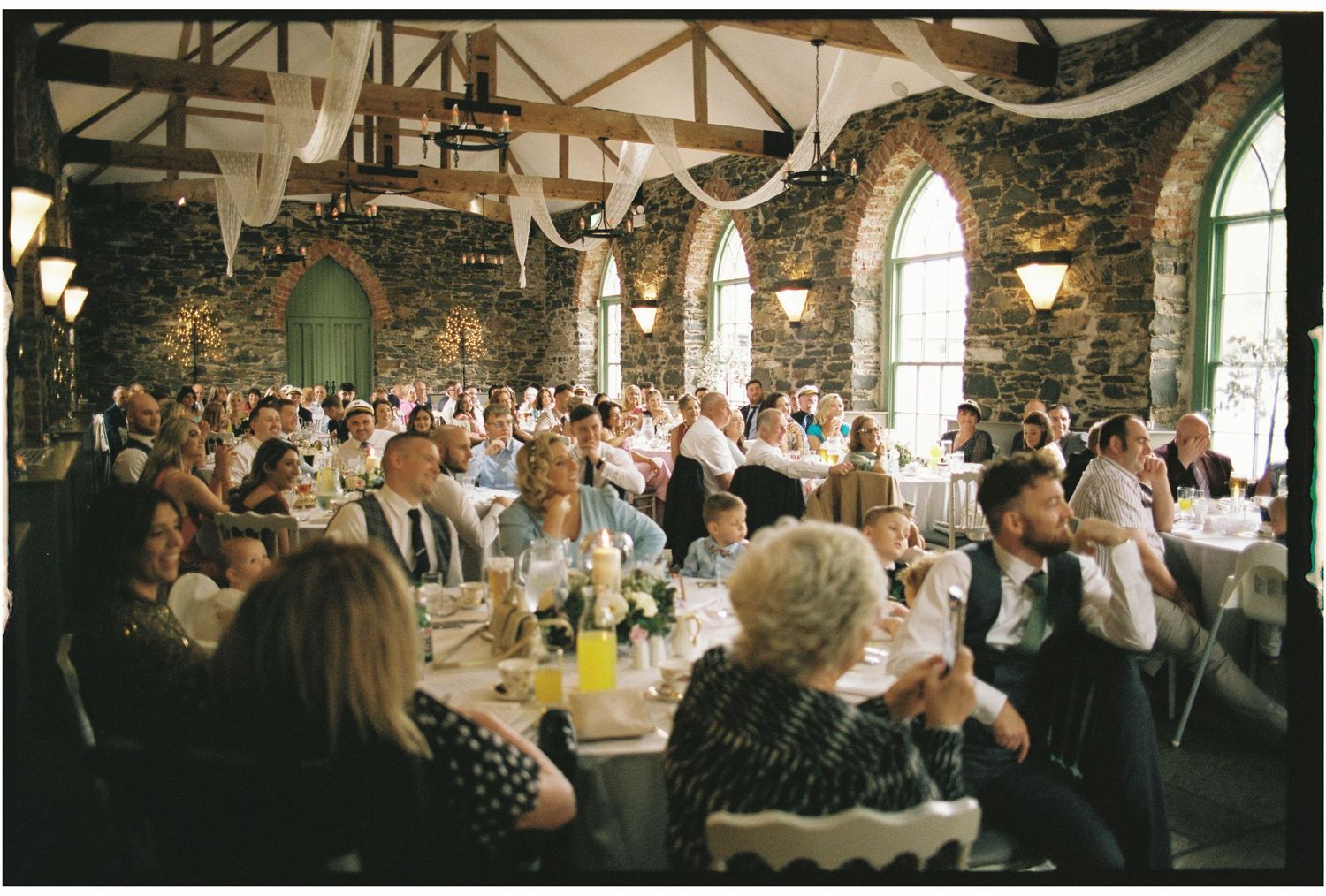 Wedding reception in a rustic stone venue with arched windows and guests seated at decorated tables.