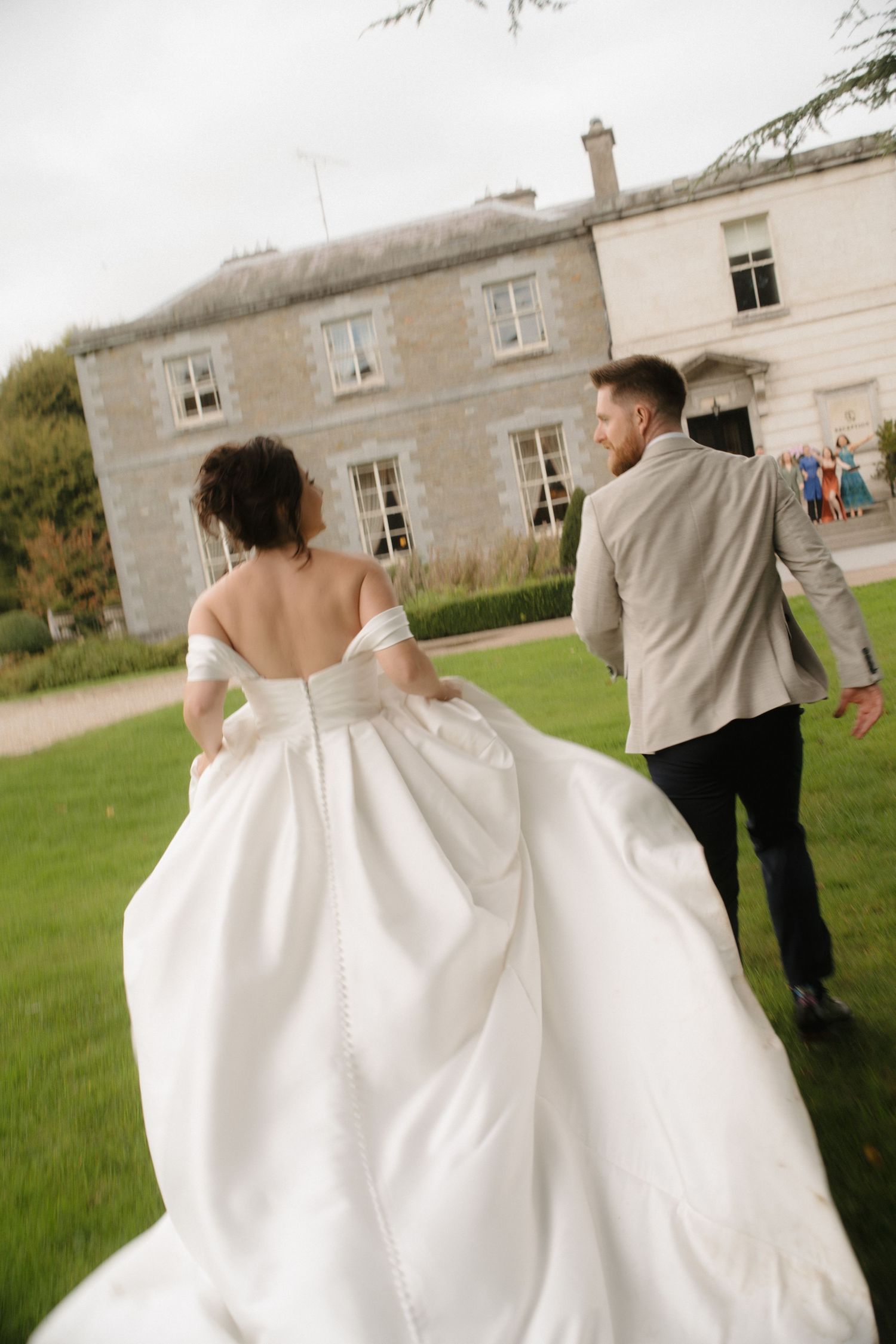 A couple in wedding attire walks across a lawn towards a grand country house with white walls.