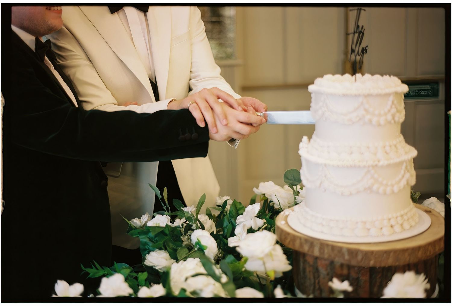 Hands in formal attire cut into a three-tiered white wedding cake decorated with flowers.
