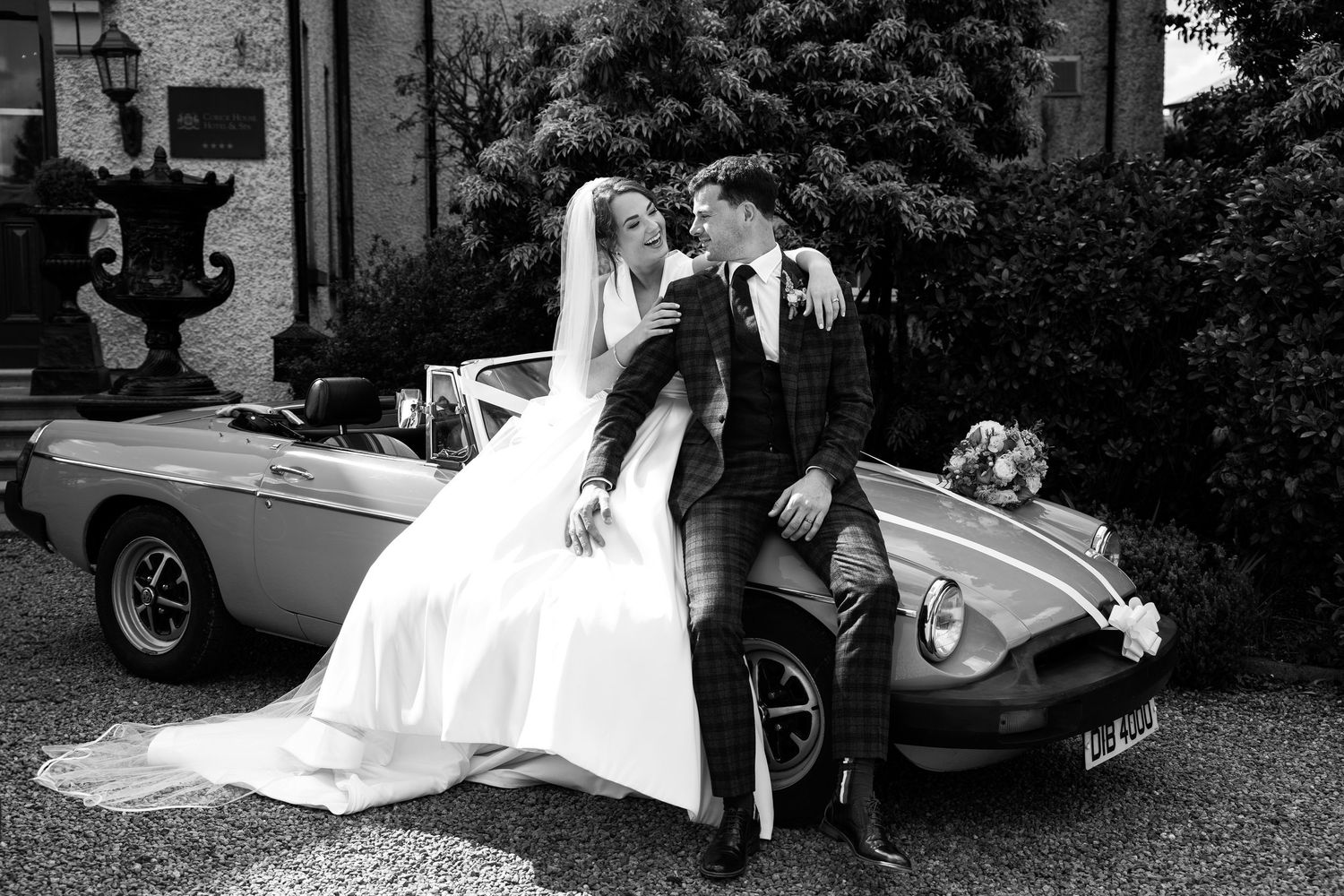 Vintage car parked outside with a bride in a flowing white wedding gown posing beside it.