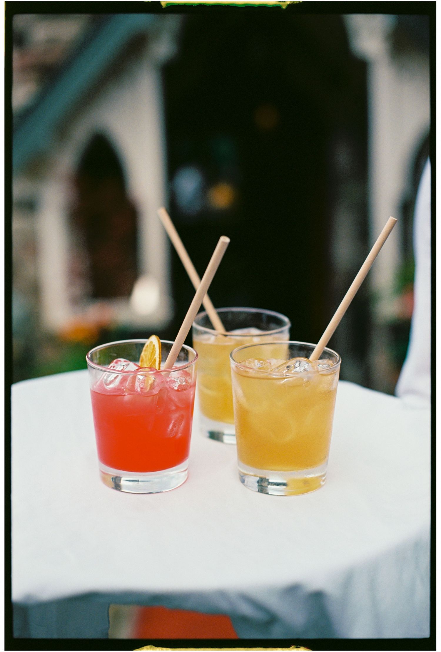 Two colorful cocktails with orange and pink hues served in rocks glasses with wooden stirrers on a white surface.