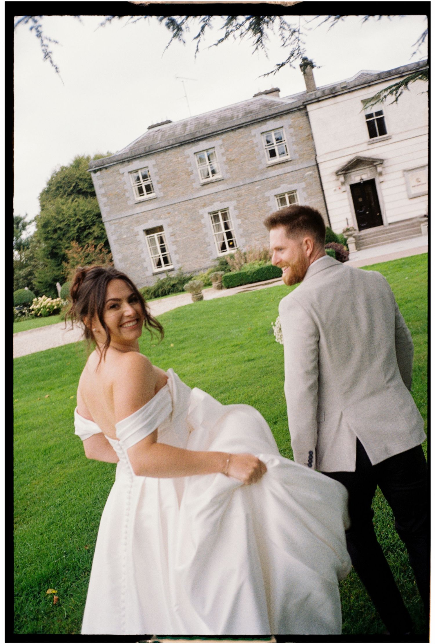 A couple in white wedding attire shares a playful moment on a manicured lawn near a stone building.