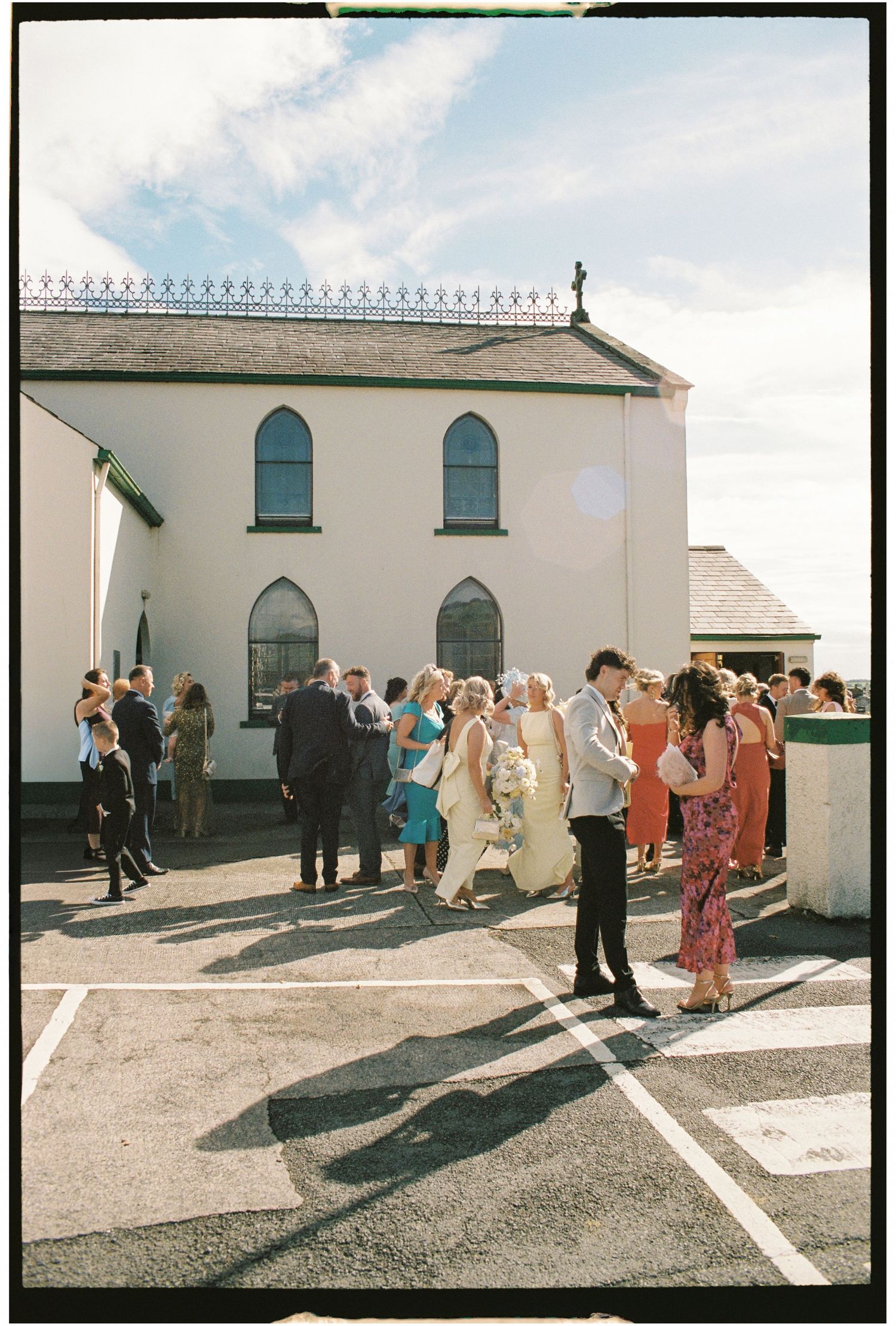 Wedding guests gather outside a white church building on a sunny day.