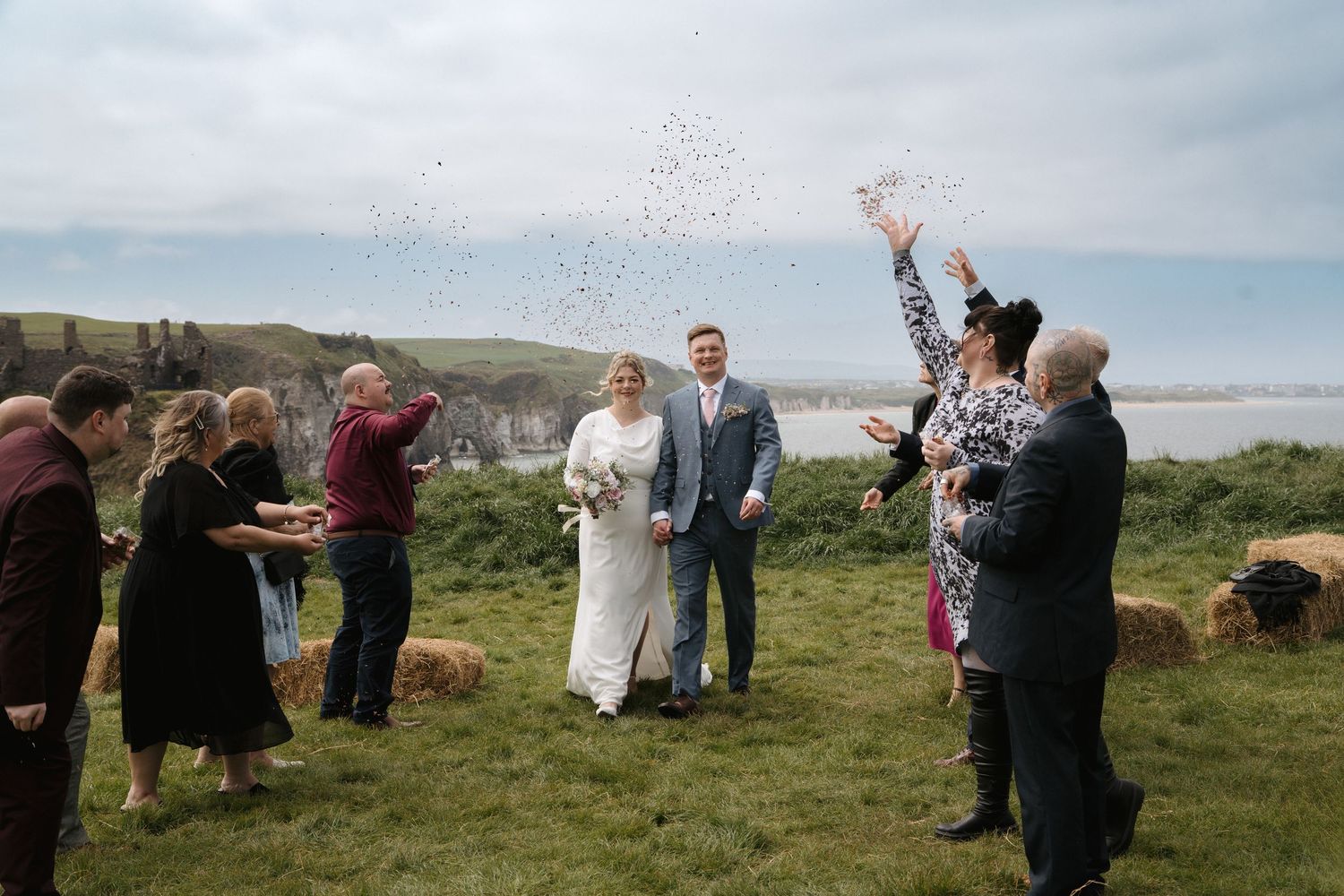 Wedding guests throw confetti over couple during outdoor coastal ceremony on grassy hilltop overlooking the ocean.