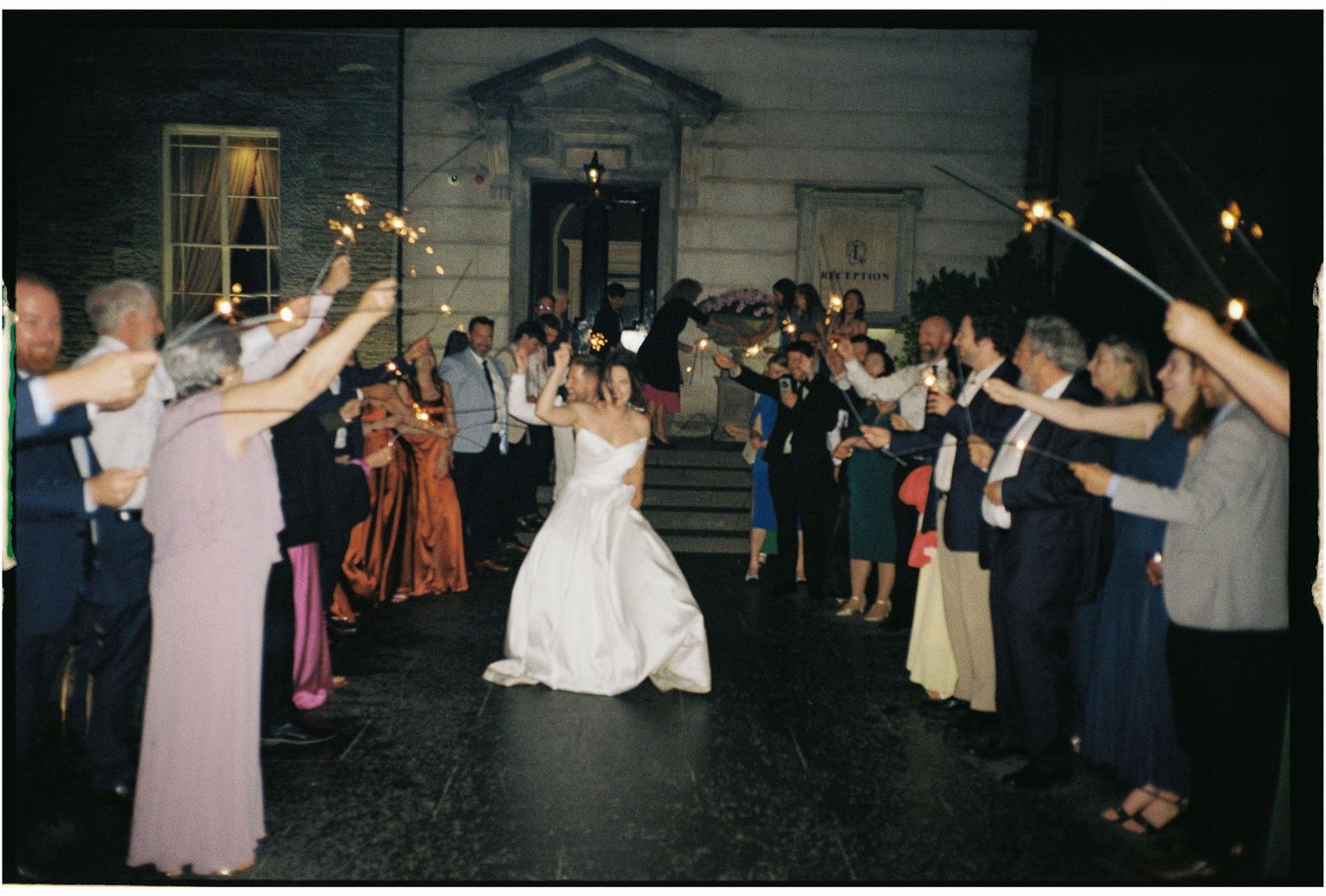 Wedding guests form a sparkler tunnel for the dancing newlyweds at an evening reception.