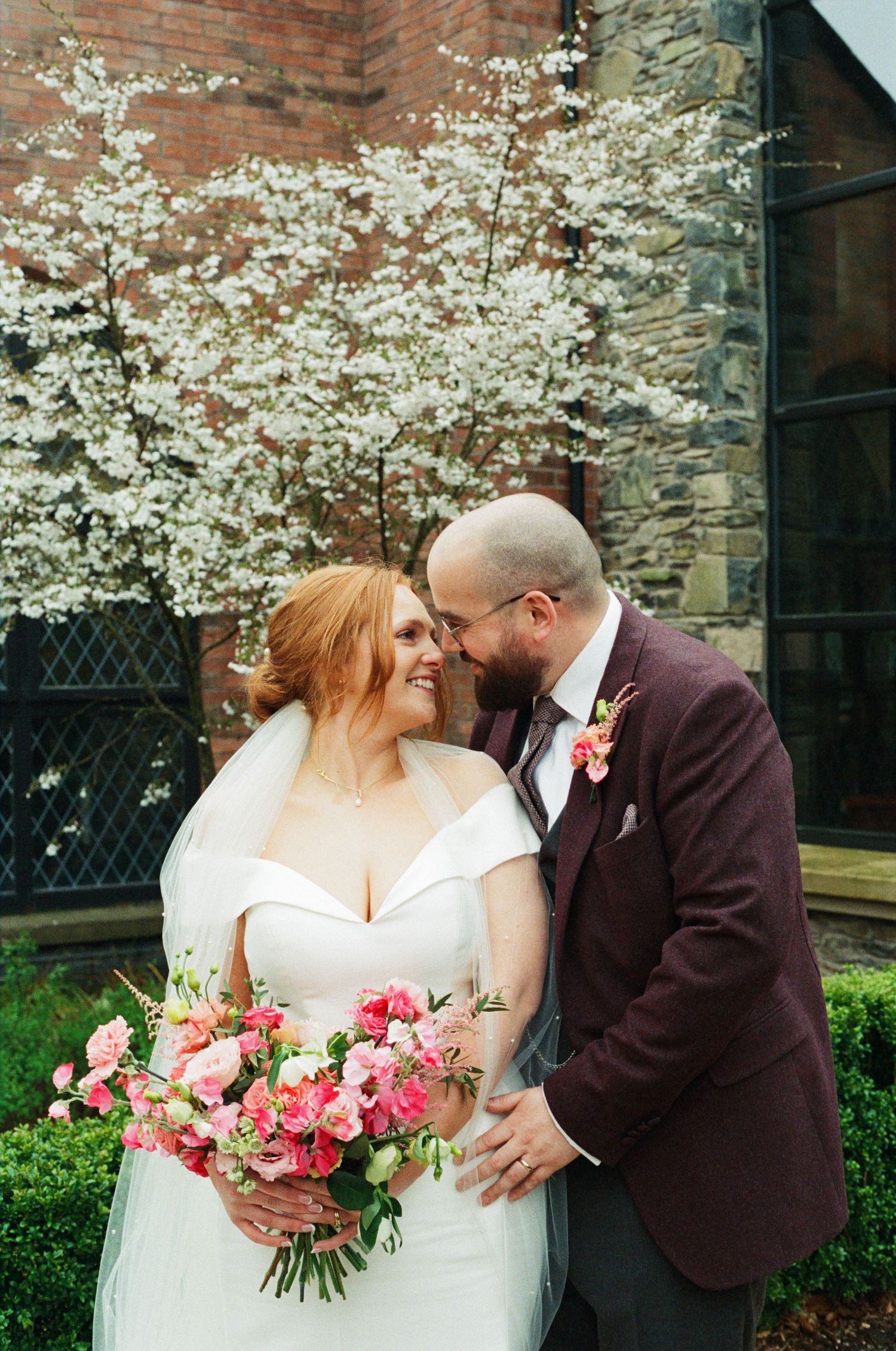 A romantic spring wedding moment with cherry blossoms blooming and a colorful pink bouquet against a stone church wall.