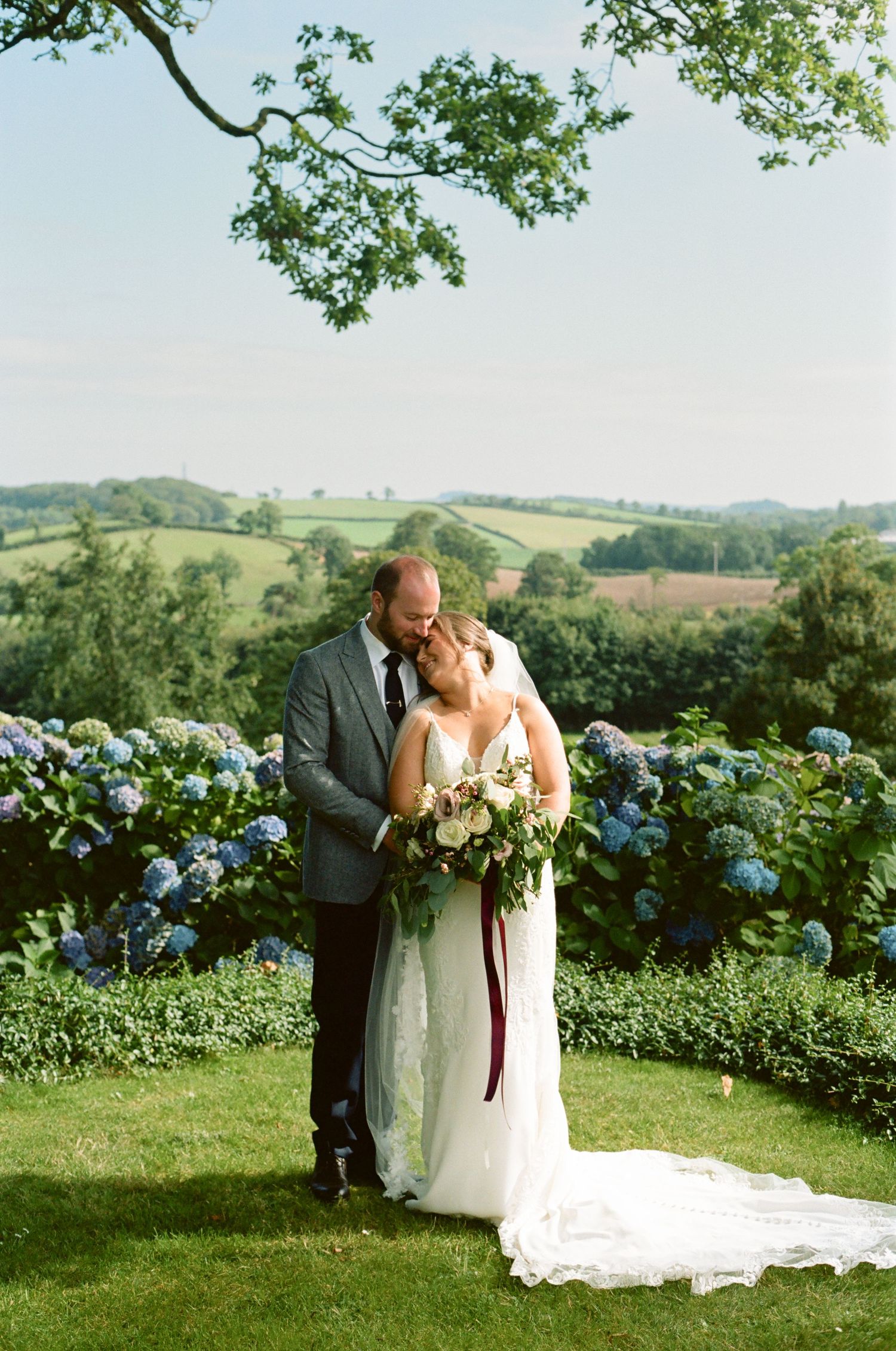 A couple shares an intimate moment together in a garden setting with rolling hills and hydrangea bushes in the background.