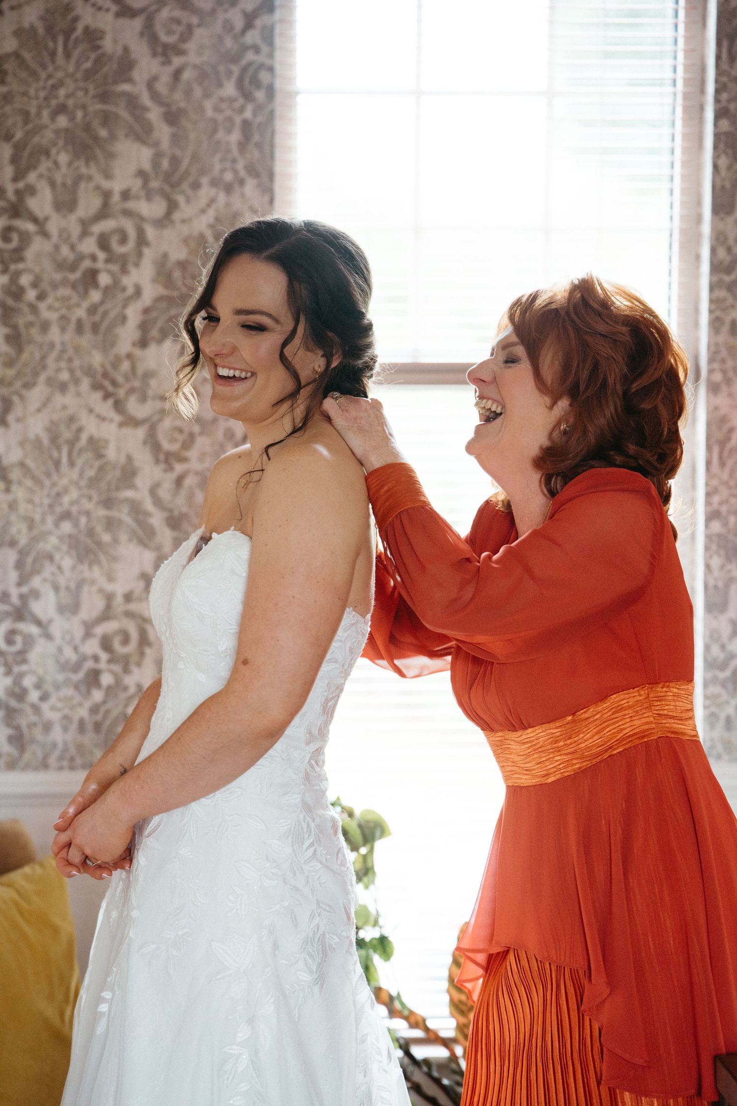 Person in orange dress helps adjust wedding gown in room with patterned wallpaper.