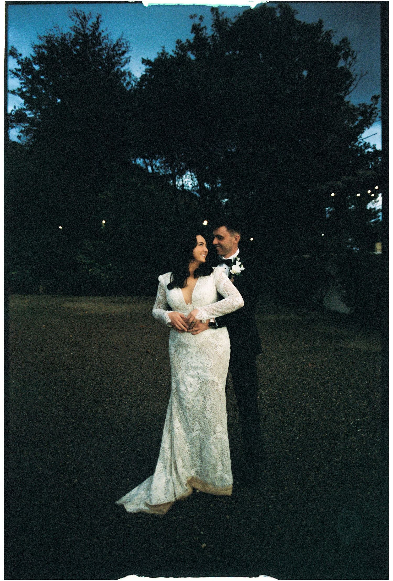 A couple dances together on a grassy lawn at dusk with trees in the background.