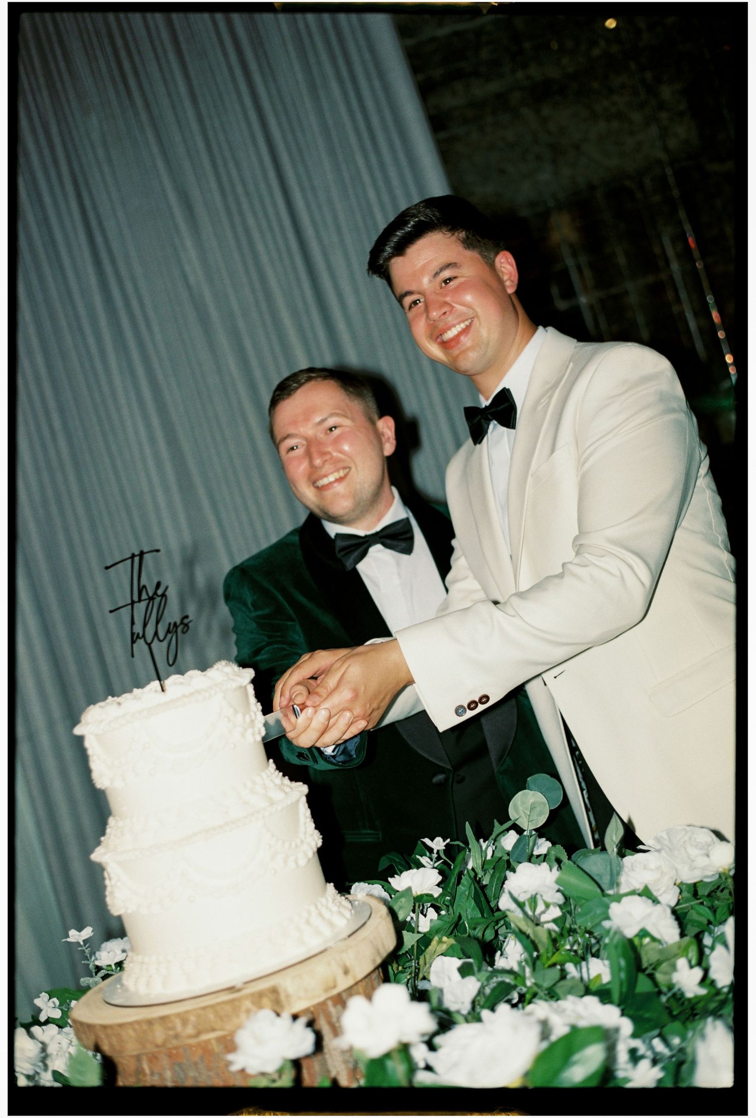 Two people in formal attire cut into a white wedding cake together surrounded by floral decorations.