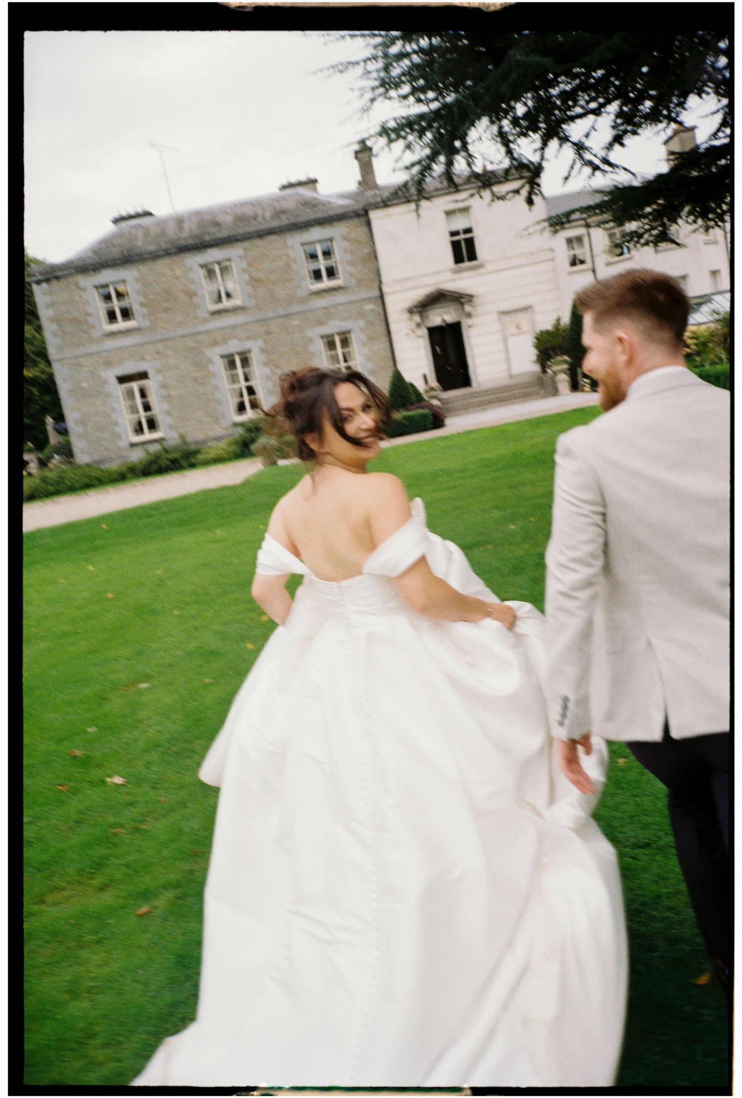 A wedding couple stands on a manicured lawn in front of an elegant stone mansion.