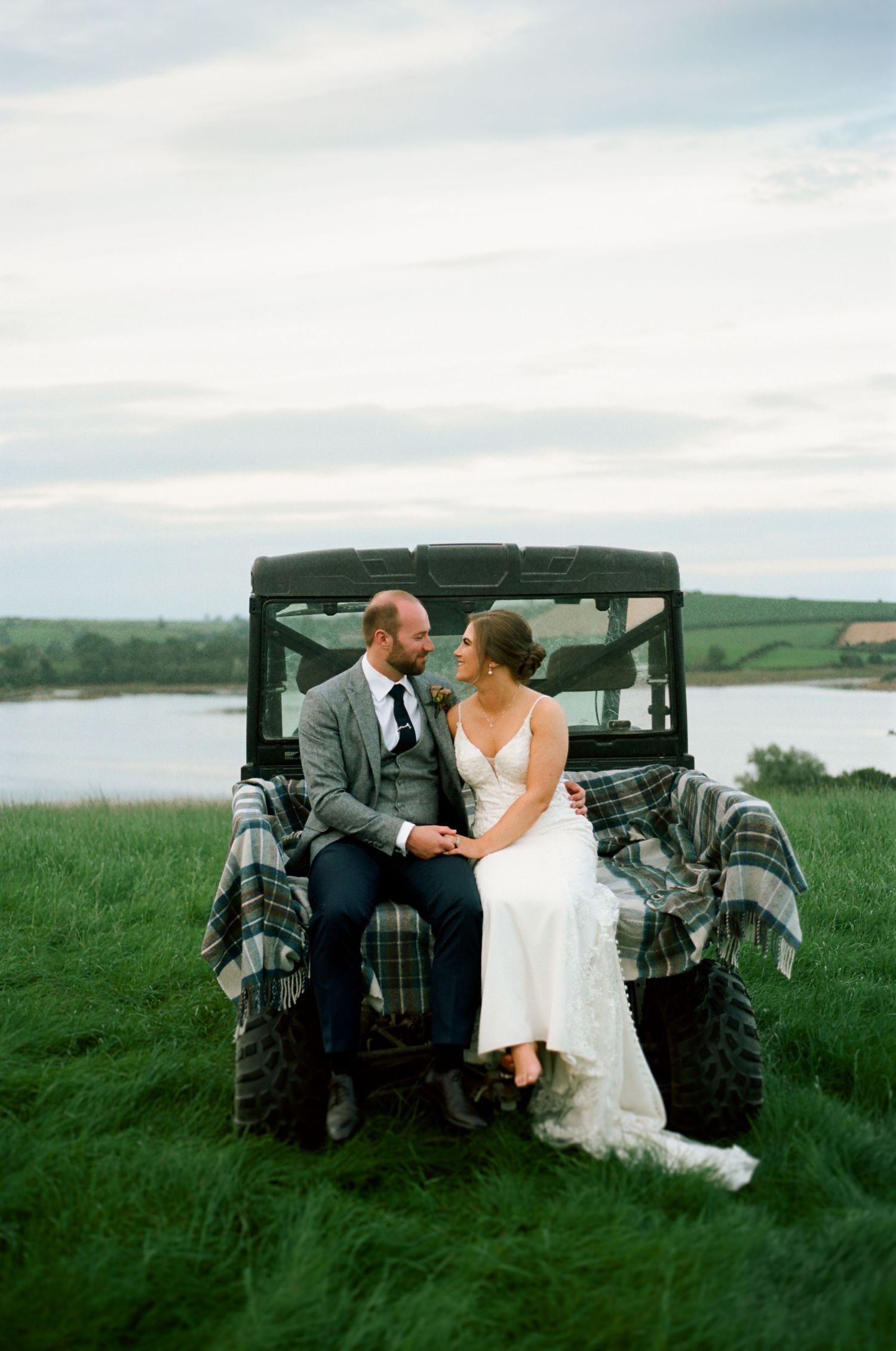 A romantic couple shares a tender moment beside a vintage car in a lush green field overlooking a serene lake.