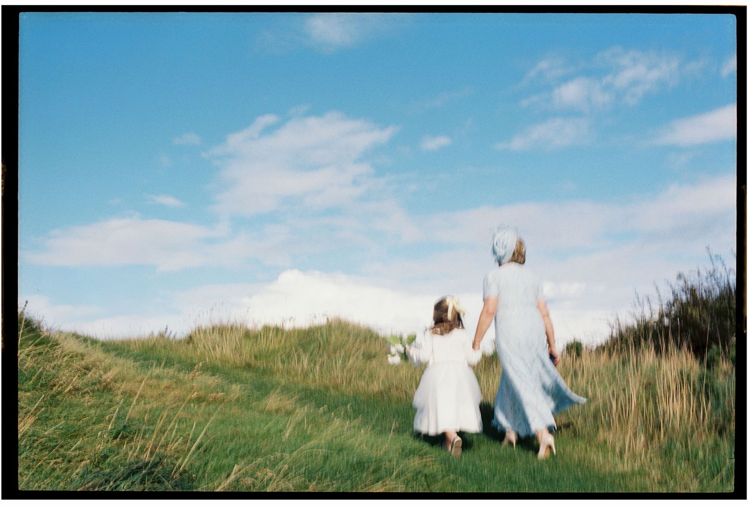 Two people in white dresses walk hand in hand through a grassy meadow under a bright blue summer sky.
