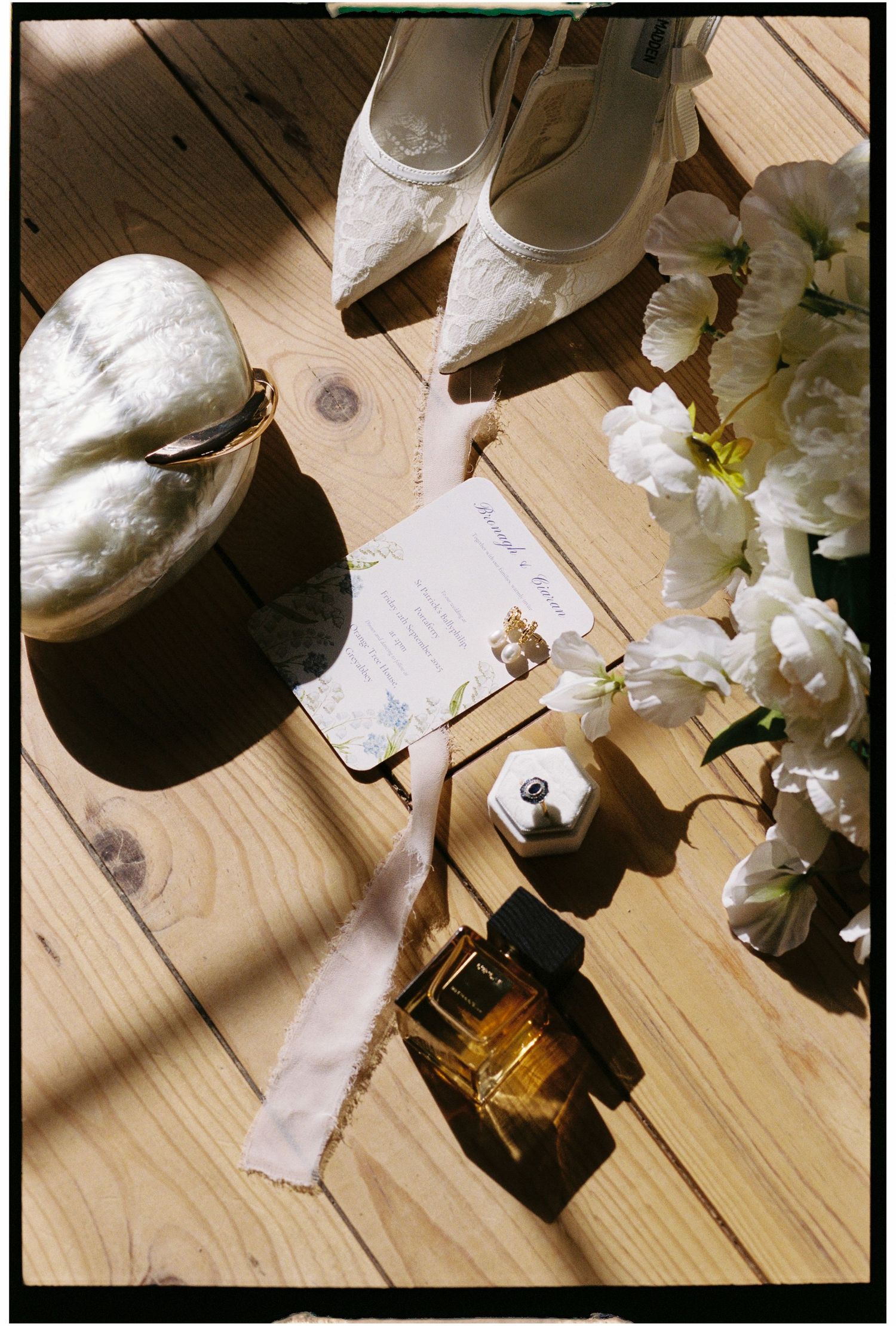 Wedding accessories including white lace shoes, perfume bottle, and white flowers arranged on wooden flooring.