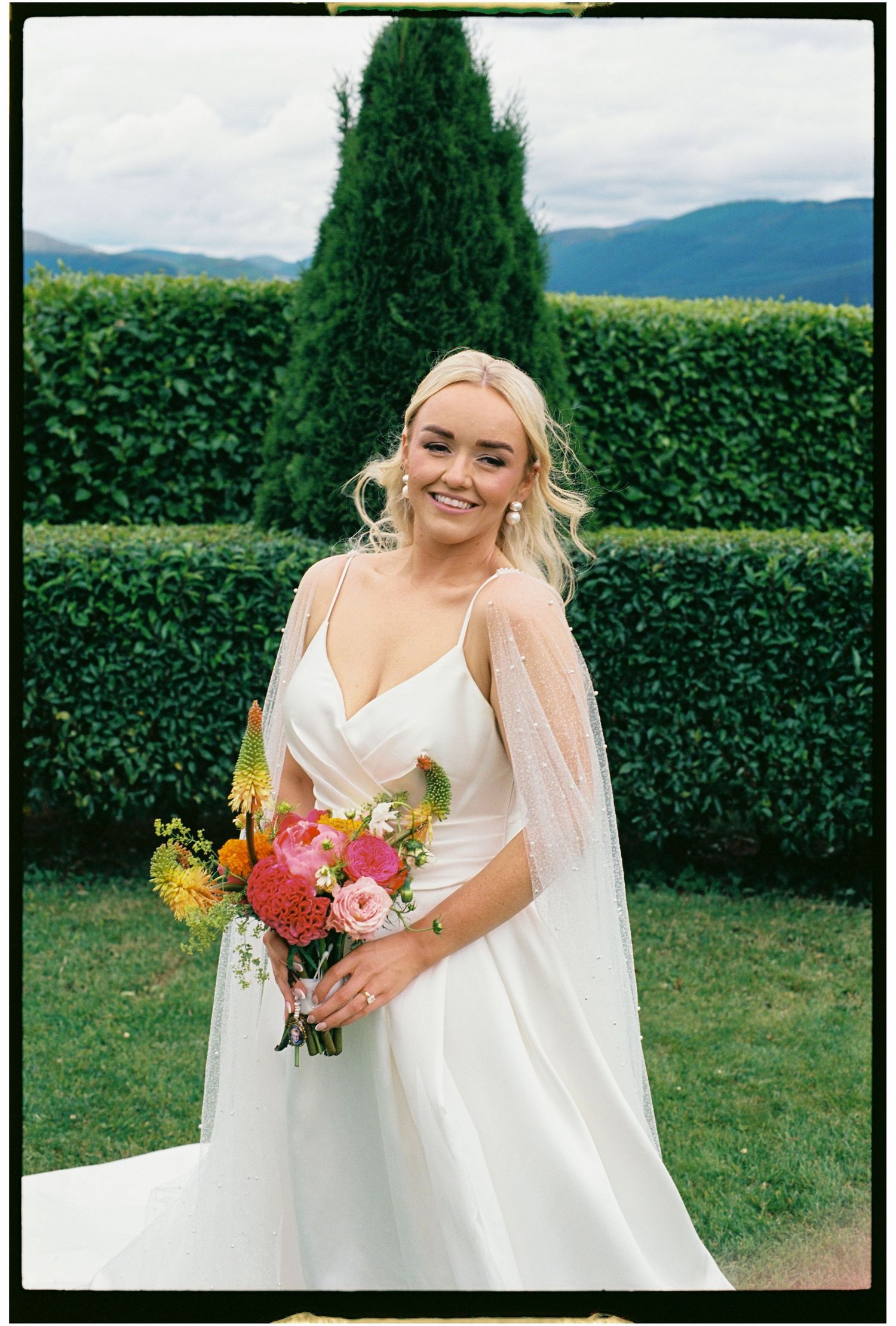 A bride in a white wedding dress poses with a vibrant bouquet against a backdrop of green hedges and mountains.