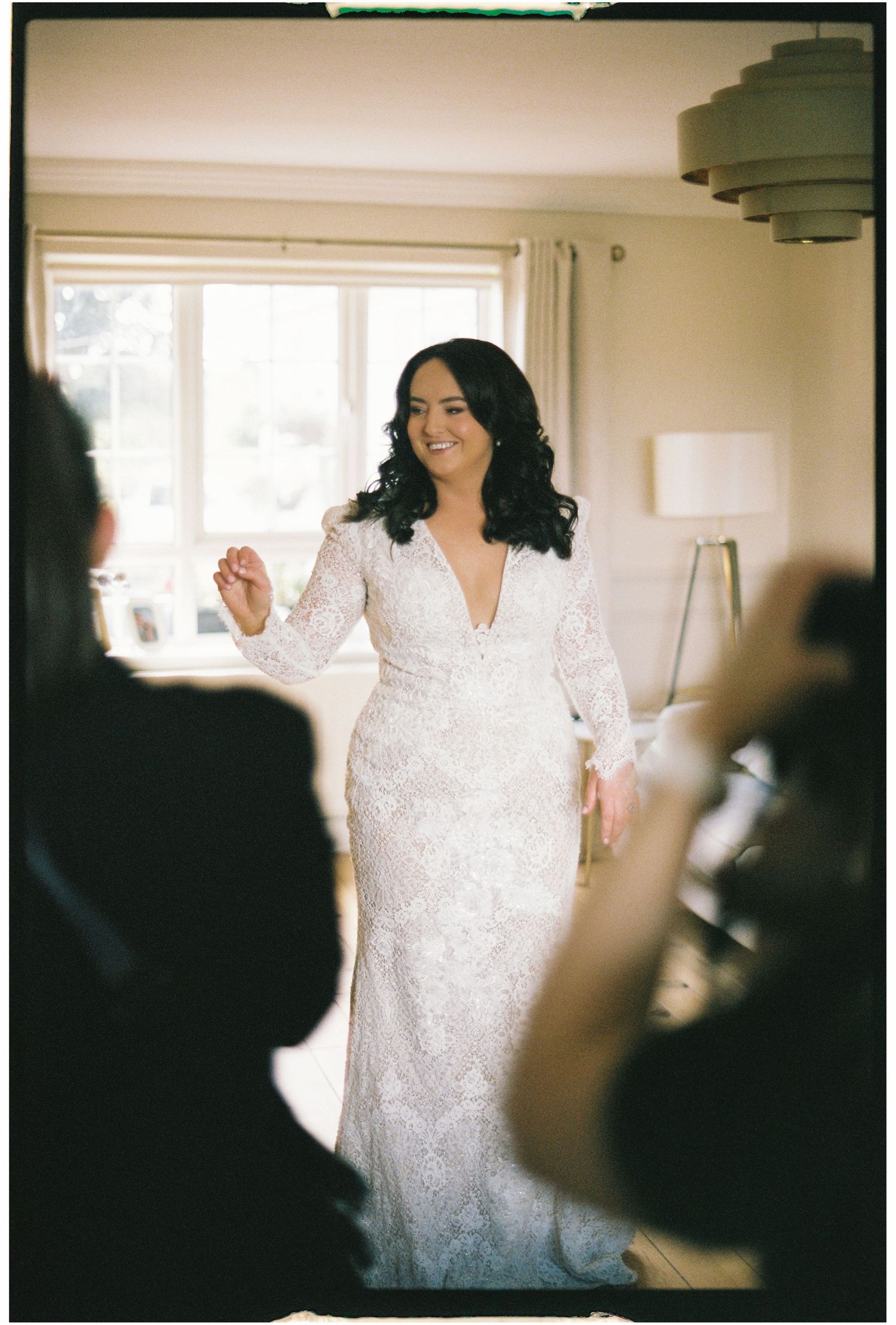 A person wearing a white lace wedding dress laughs joyfully while getting ready in a sunlit room.