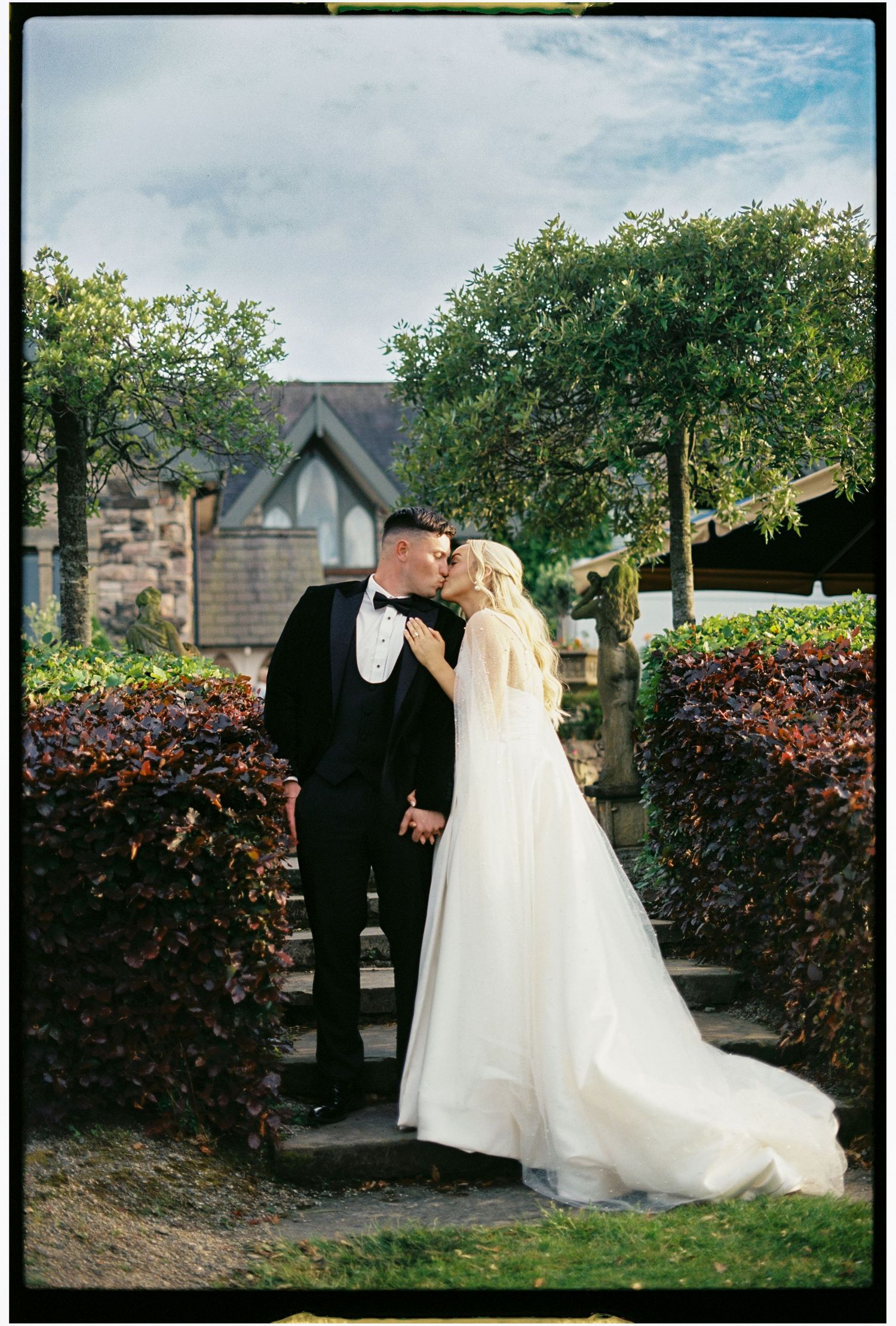 A wedding couple shares a tender moment in a garden setting with lush hedges and stone architecture.